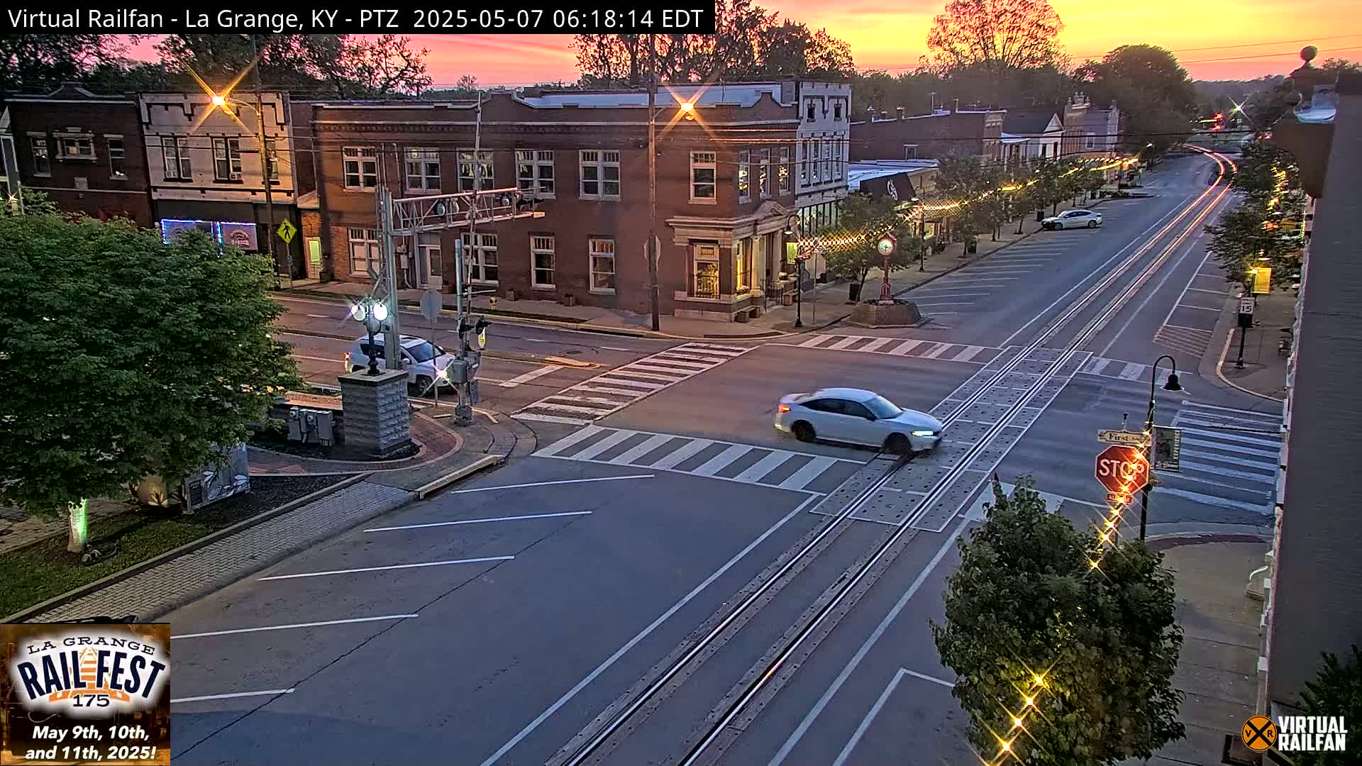 A quiet, sun-drenched street scene at sunrise shows a car driving through a town square with brick buildings on either side of the road.