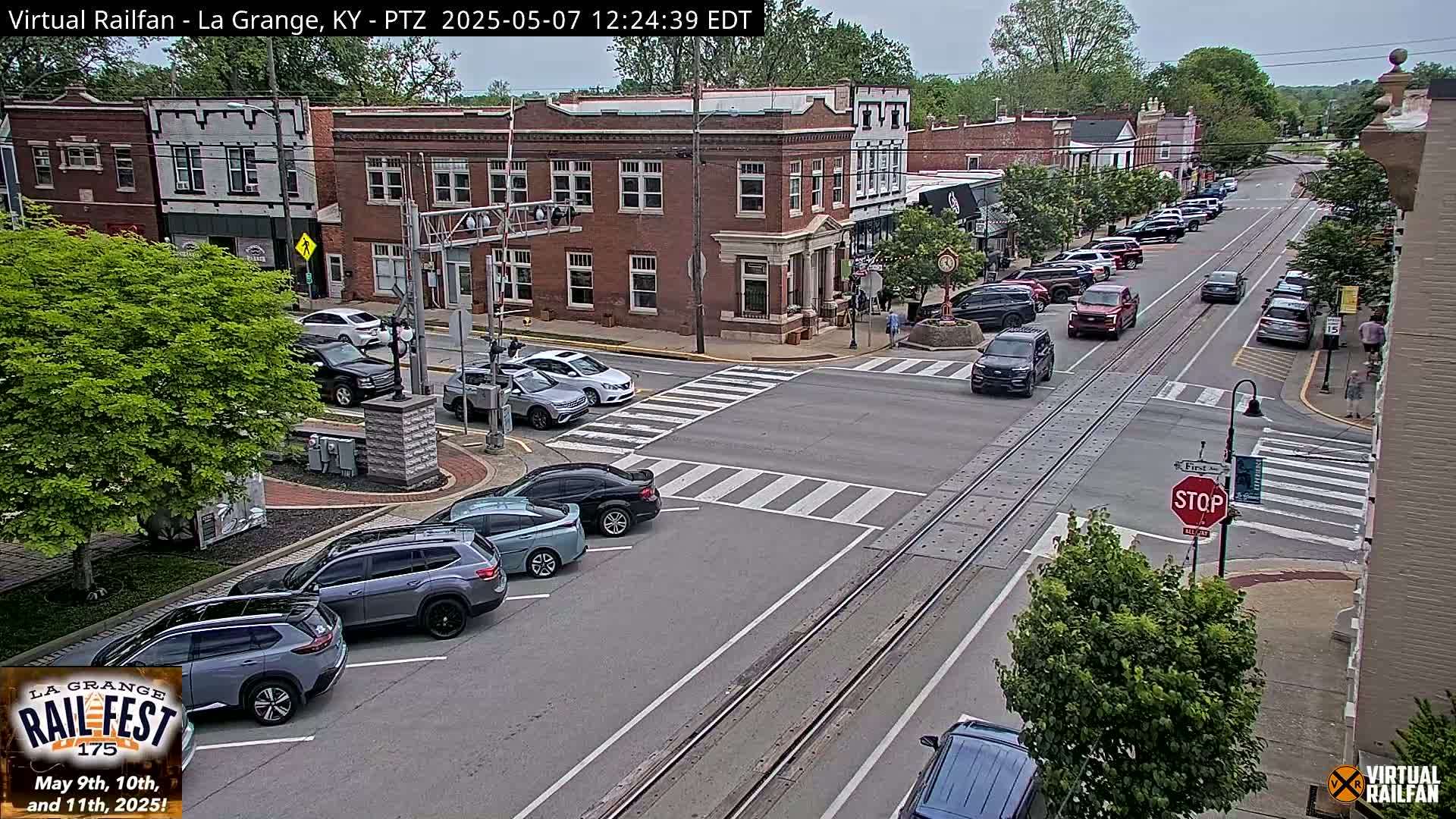 A partly sunny day in a small town features a street intersection with several cars, brick buildings, and lush green trees.