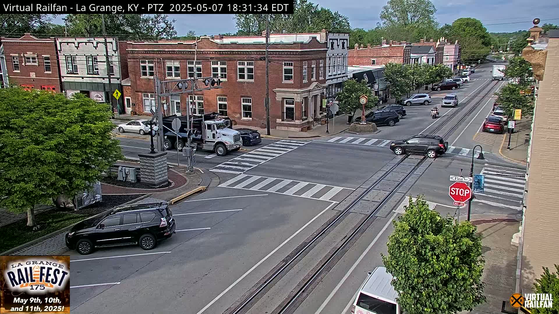 A mostly sunny view of a town's street intersection with brick buildings, parked cars, and a few vehicles in motion, featuring train tracks running down the center of the road.