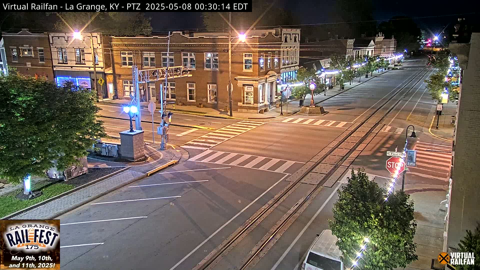 A nighttime street scene shows a brick building at an intersection with train tracks, streetlights, and crosswalks.