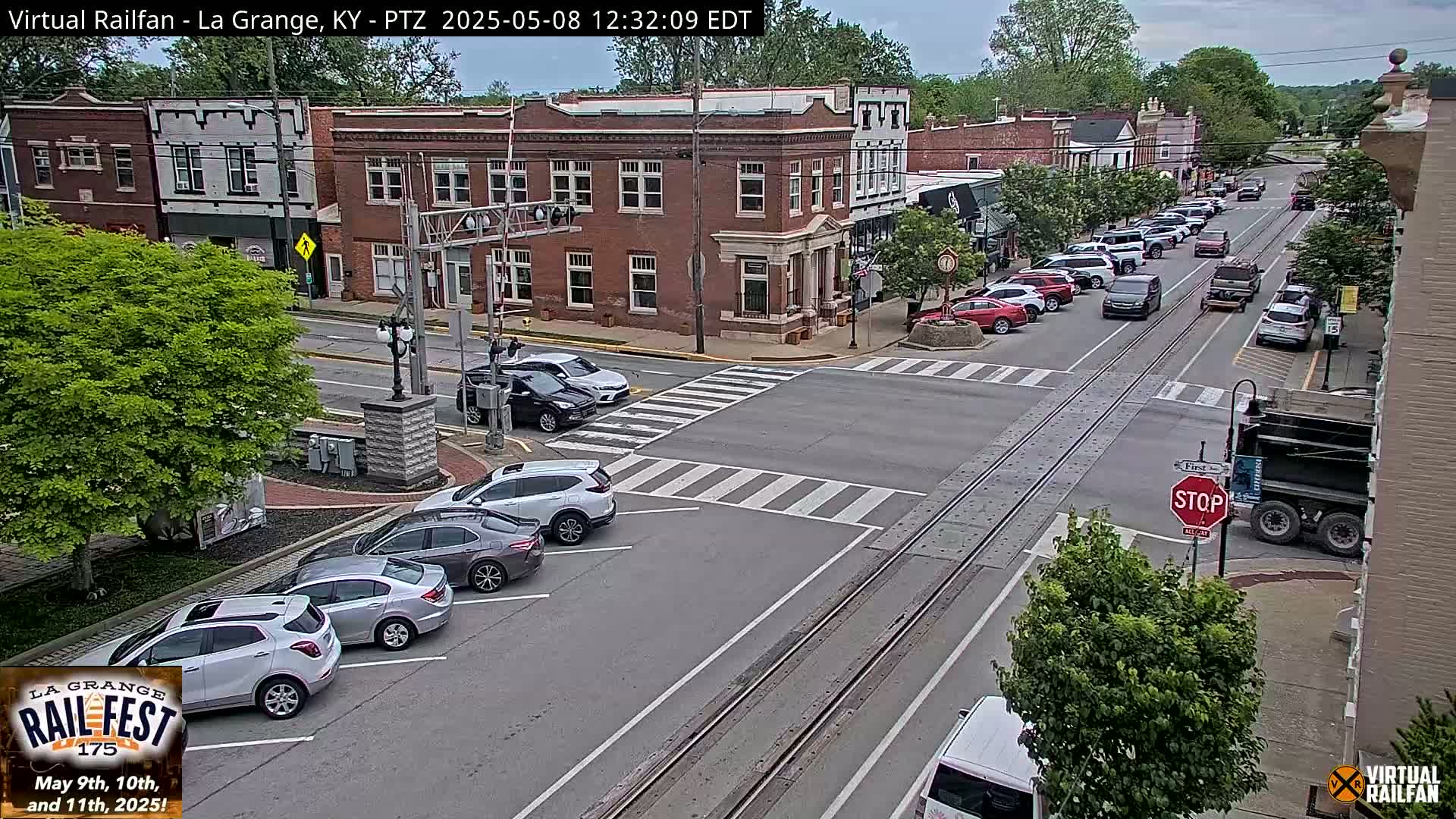 A mostly sunny street scene in a small town shows several cars parked along the street and several more driving on the road, which has train tracks running down the center.