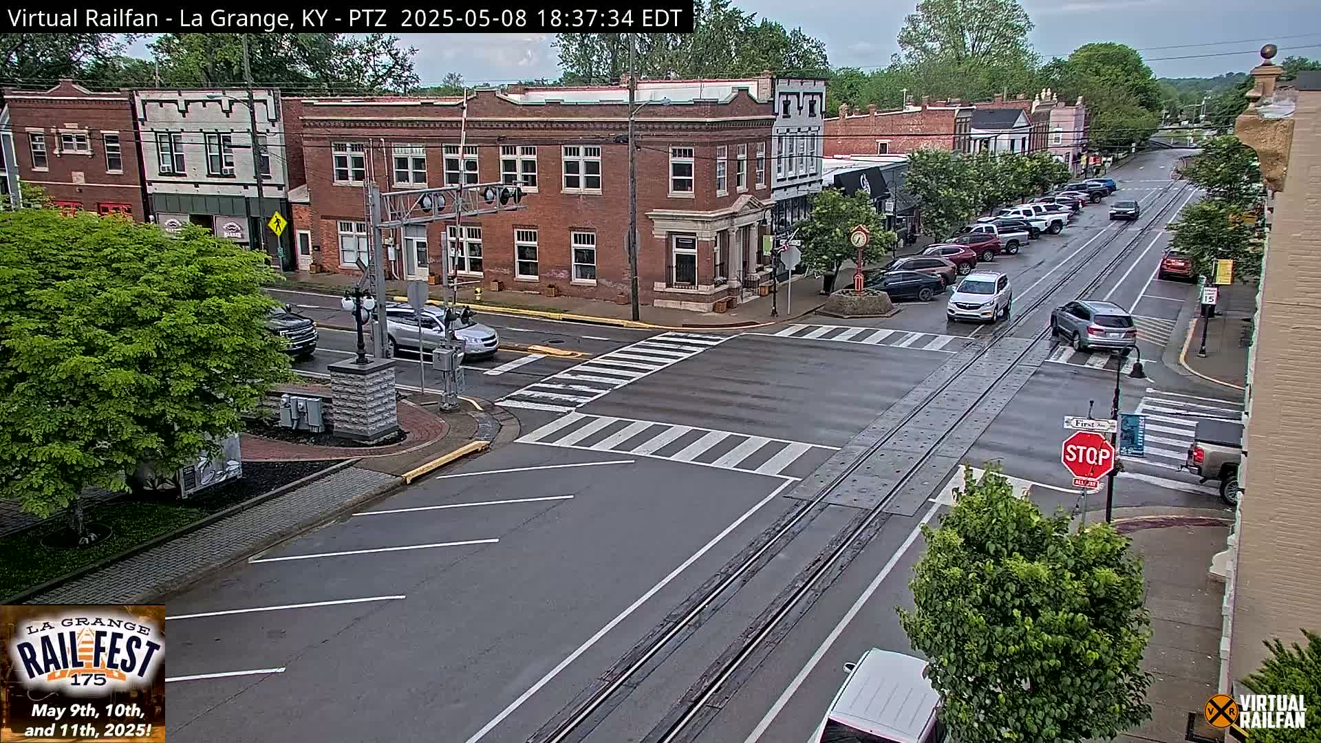 A wet street scene shows a town square with several cars at an intersection, brick buildings, and lush green trees under an overcast sky.