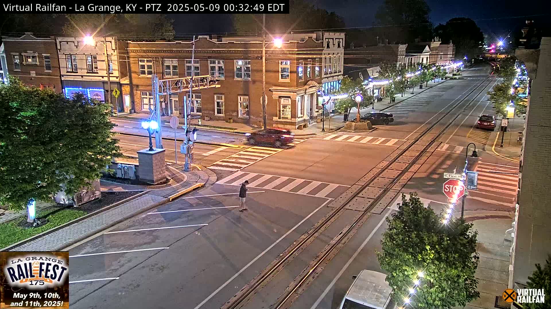 A nighttime street scene shows a person standing on a crosswalk at an intersection with train tracks, near brick buildings and streetlights.
