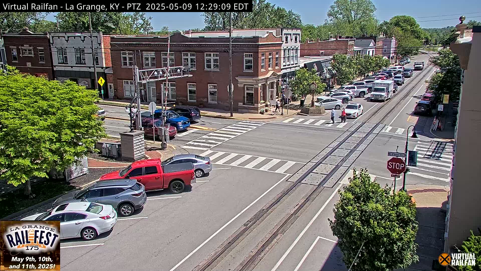 A sunny day in a town square, showing several parked and moving vehicles at a street intersection with train tracks, brick buildings and lush green trees.