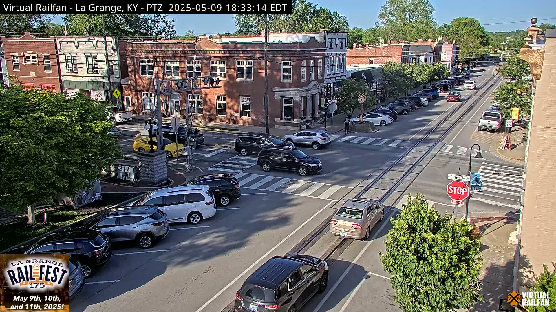 A sunny town square with several cars parked and driving on the street, intersecting train tracks, and brick buildings.