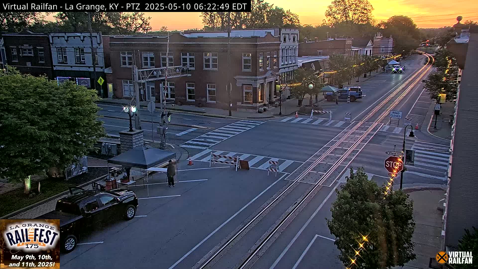 A mostly empty street scene at sunset shows a town square with brick buildings, a few vehicles, and a person standing near a tent.