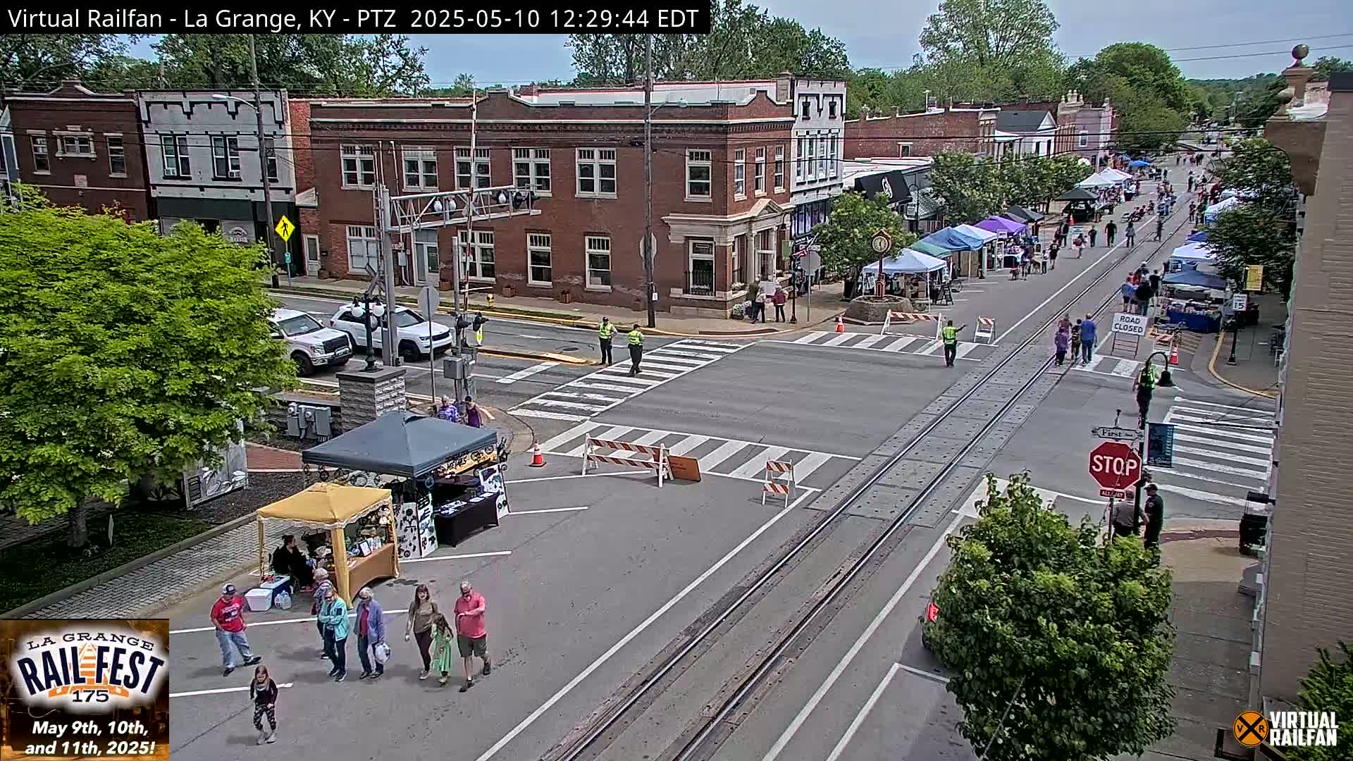 A street scene on a sunny day shows a town with vendors' tents set up along a road closed for an outdoor event, with people walking around and several cars parked along the sides of the road.