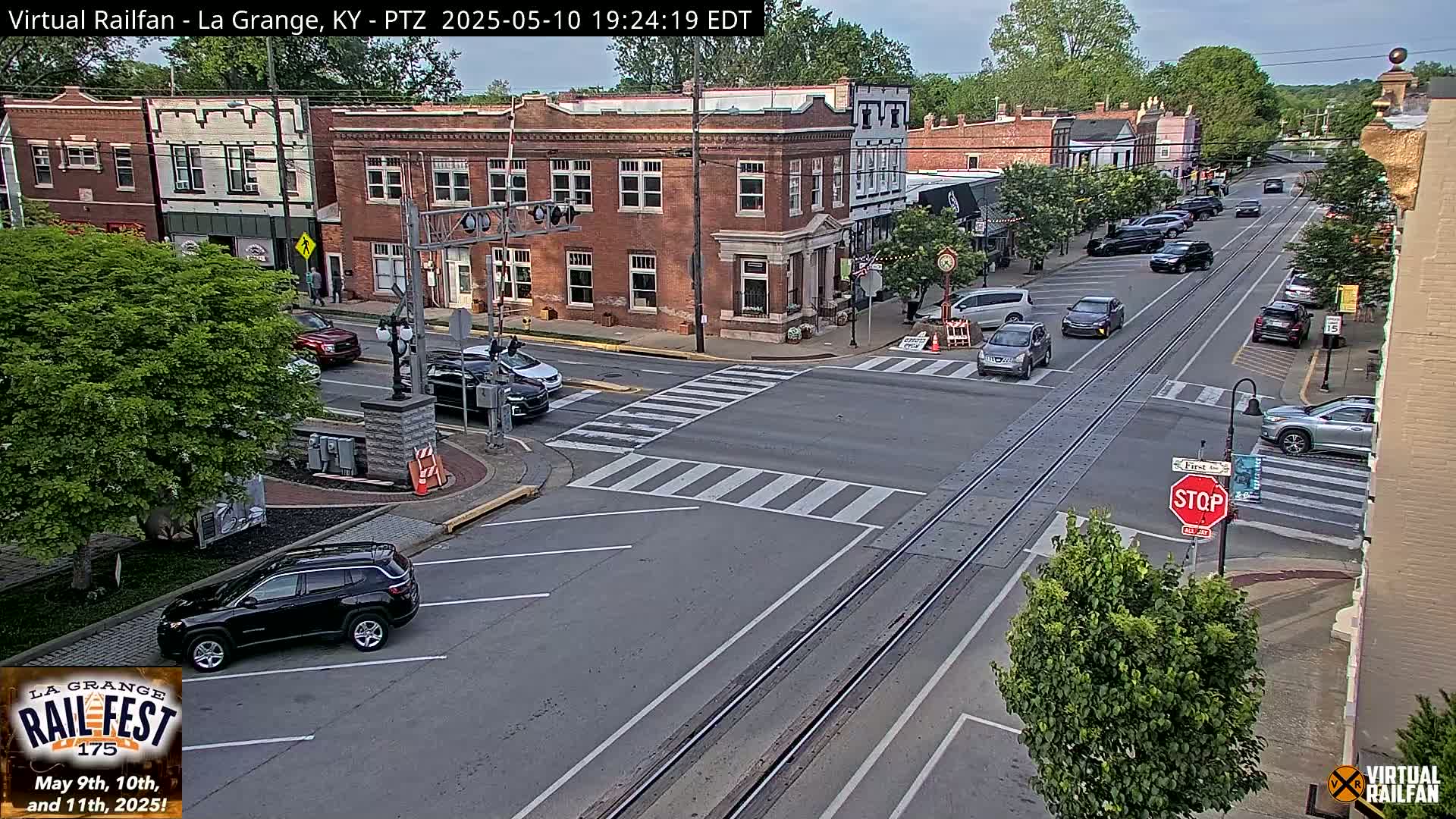 A partly sunny day reveals a town square with several cars at an intersection, brick buildings lining the street, and train tracks running through the center.