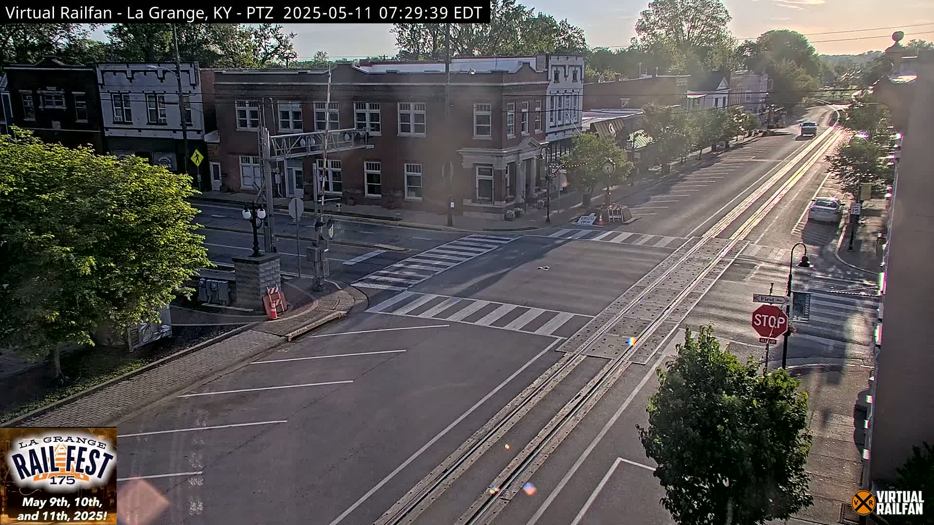 A sunny street scene shows a two-lane road with train tracks running down the center, passing through a town with brick buildings and green trees.