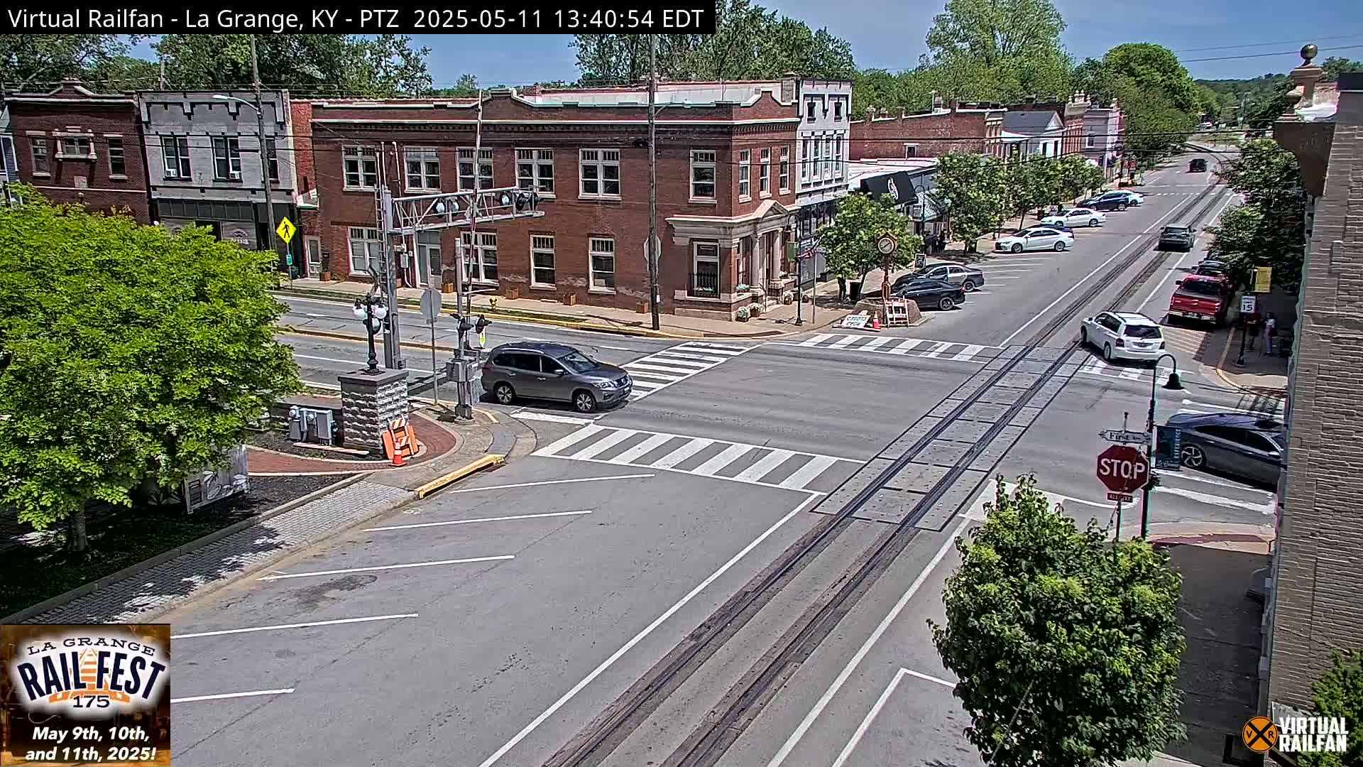 A sunny day shows a view of a town's intersection with parked cars and a few cars driving, railroad tracks crossing the road, and brick buildings lining the street.