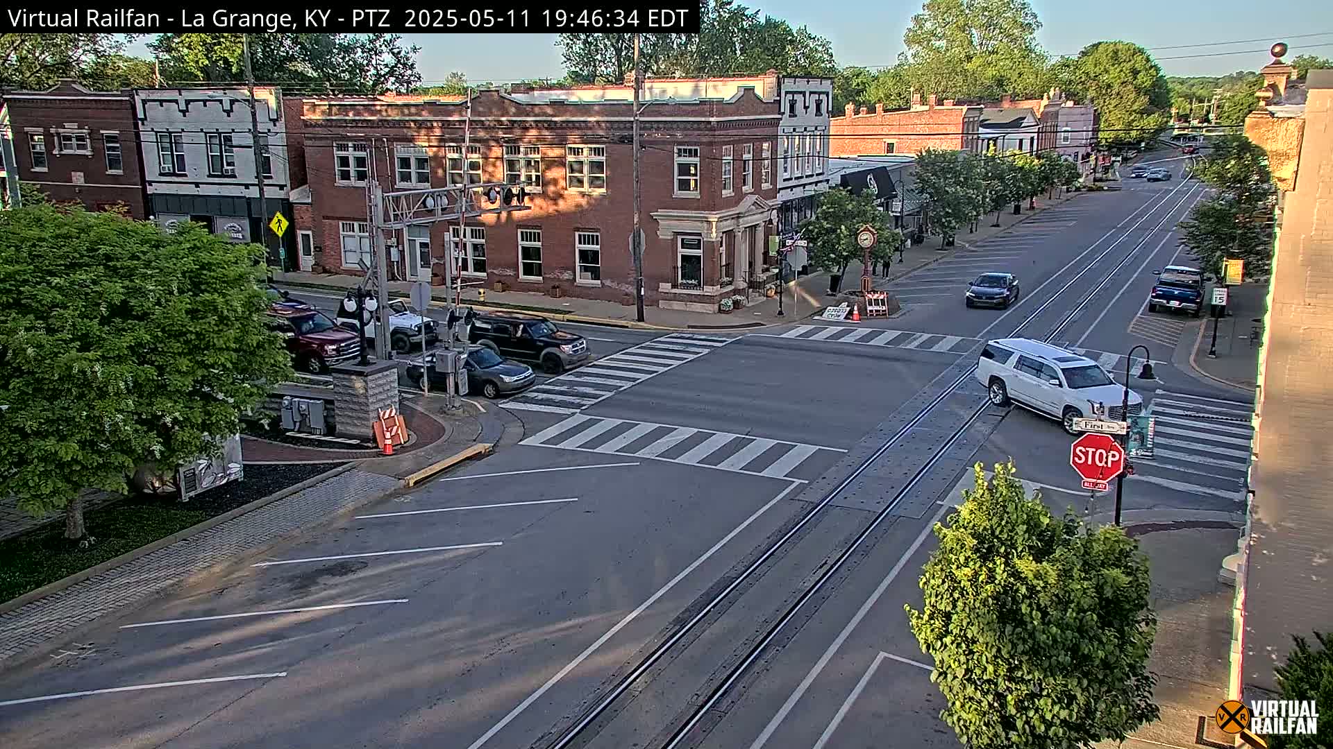A sunny daytime view of a street intersection in a small town, showing brick buildings, cars, and a railway crossing.
