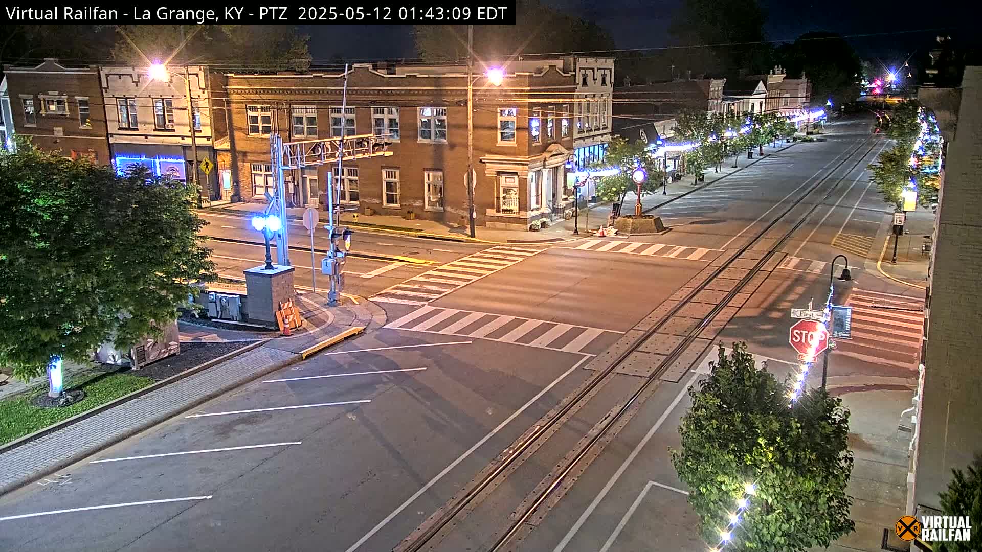 A nighttime view of a quiet street intersection with train tracks, streetlights, and buildings illuminated.