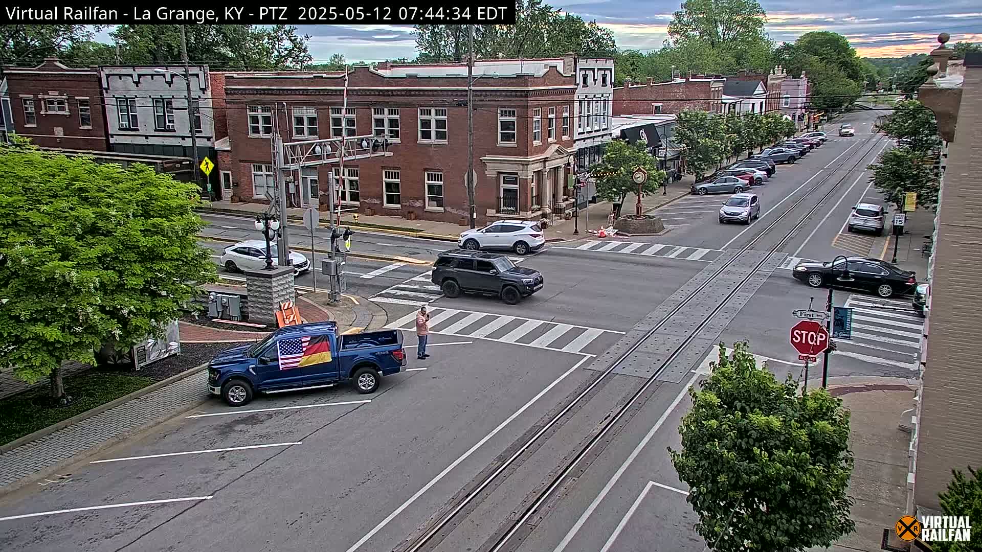 A mostly sunny view of a town intersection with several cars and a person standing near a blue pickup truck displaying American and German flags.