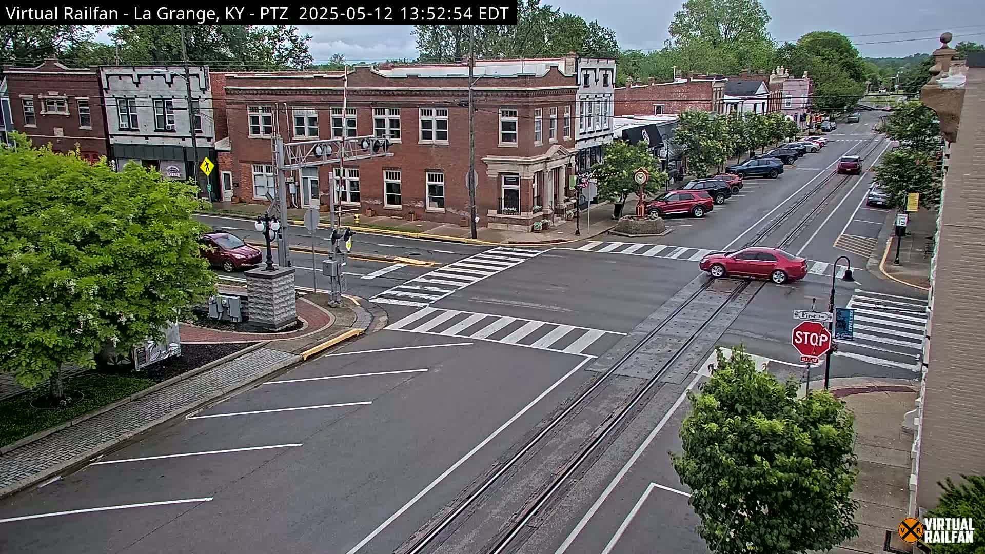 A mostly overcast view of a town square intersection with brick buildings, parked cars, and a red car crossing train tracks.