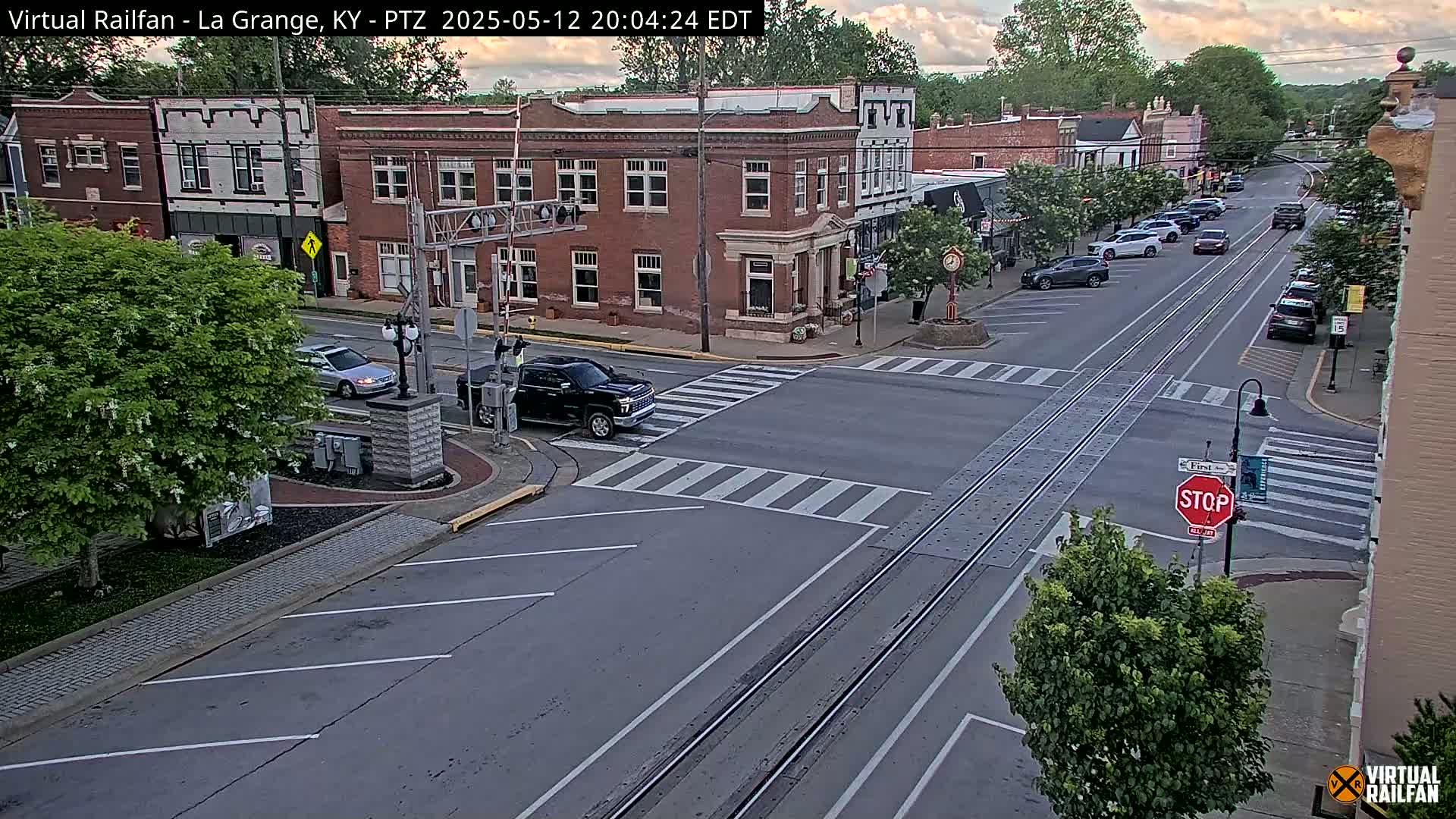 A street scene at dusk shows a town square with brick buildings, cars parked along the sides of the street, and a few cars moving through the intersection.