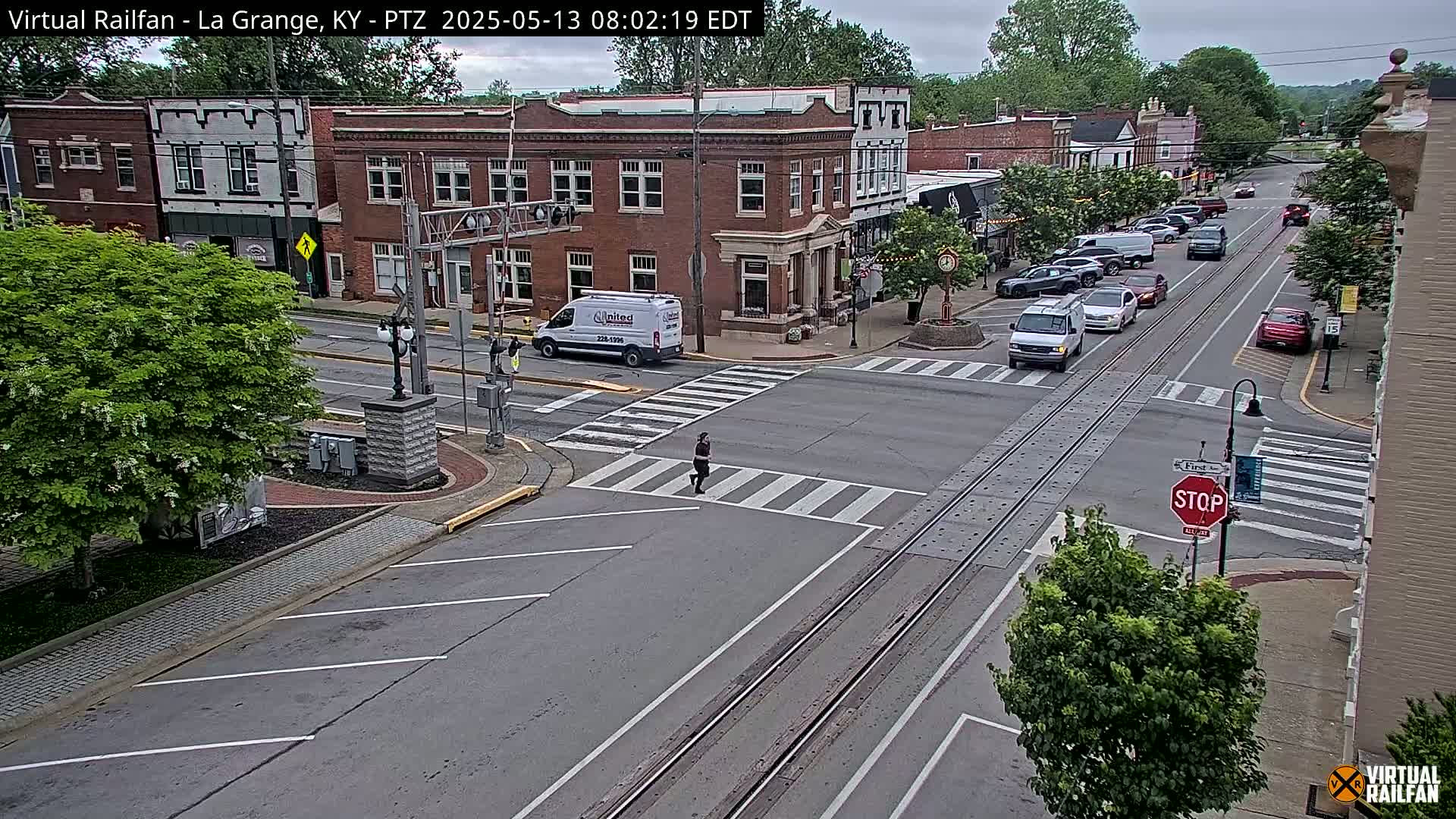 A mostly sunny day in a small town shows a street intersection with several cars and a pedestrian crossing railroad tracks.