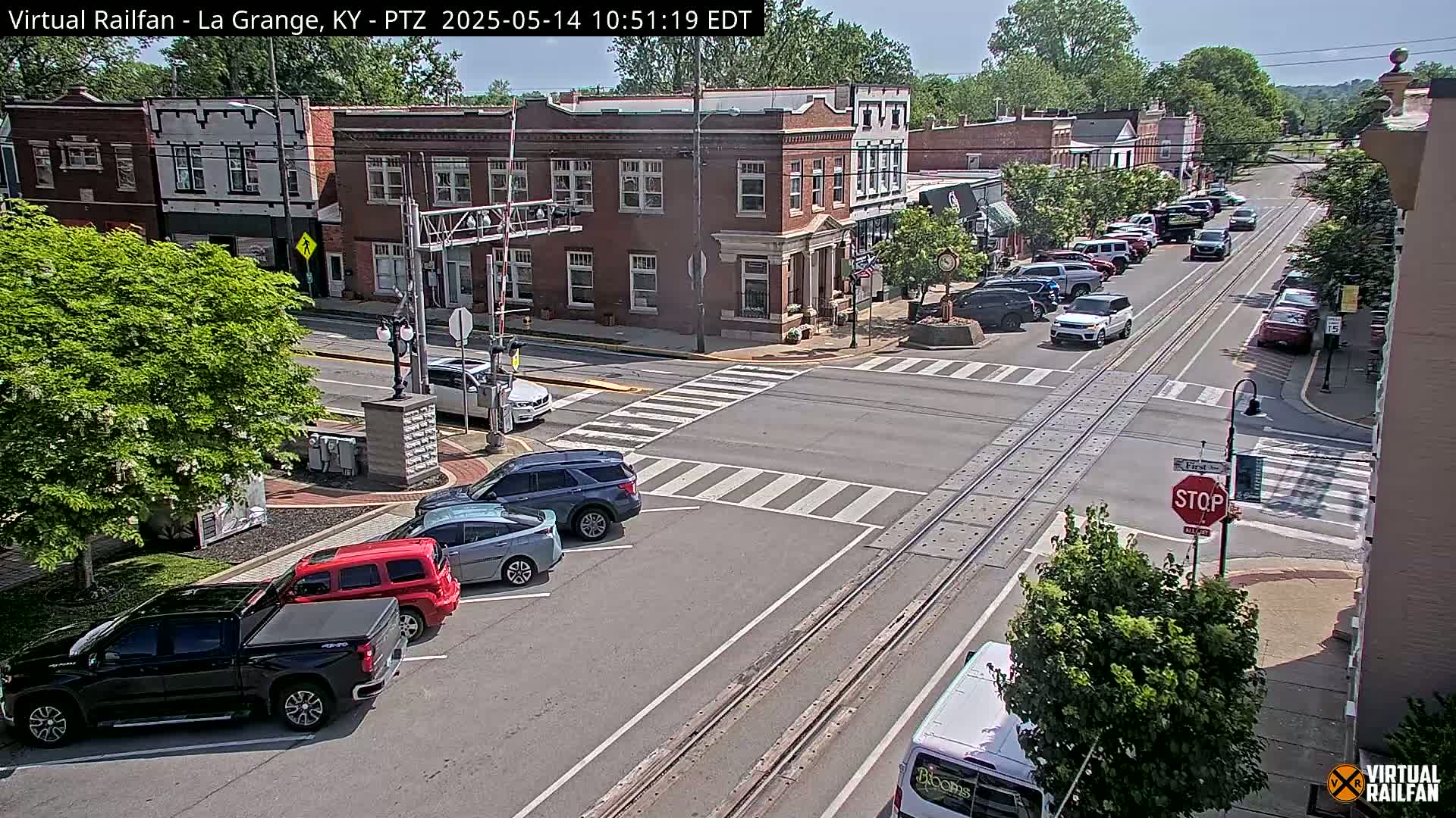 A sunny day view of a town's intersection with several cars parked and driving along streets that intersect with train tracks.