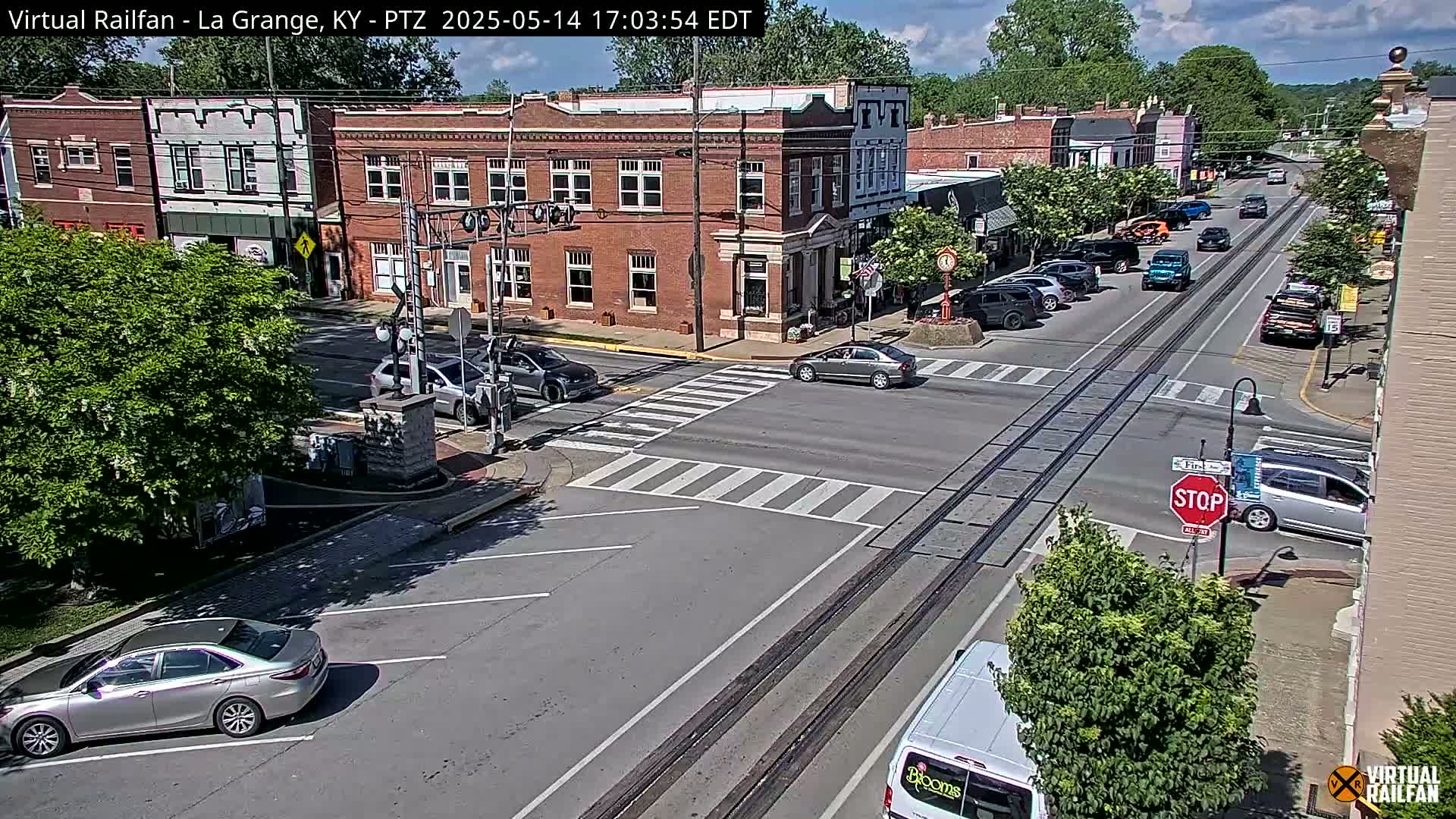 A sunny street scene in a small town shows several cars at an intersection with railroad tracks running down the middle of the road, bordered by brick buildings and lush green trees.