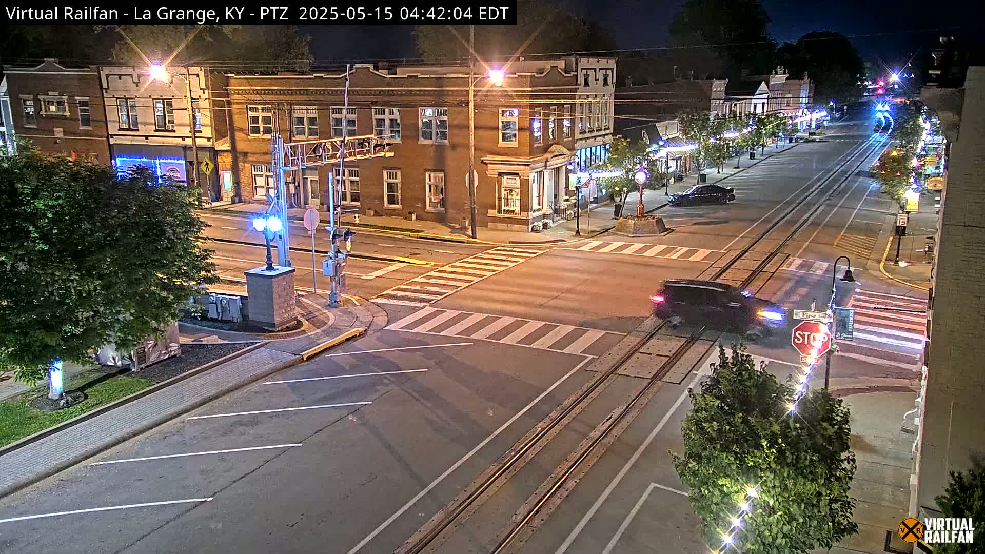 A nighttime street scene shows a vehicle crossing a railroad track at a crosswalk in front of brick buildings with streetlights illuminating the area.