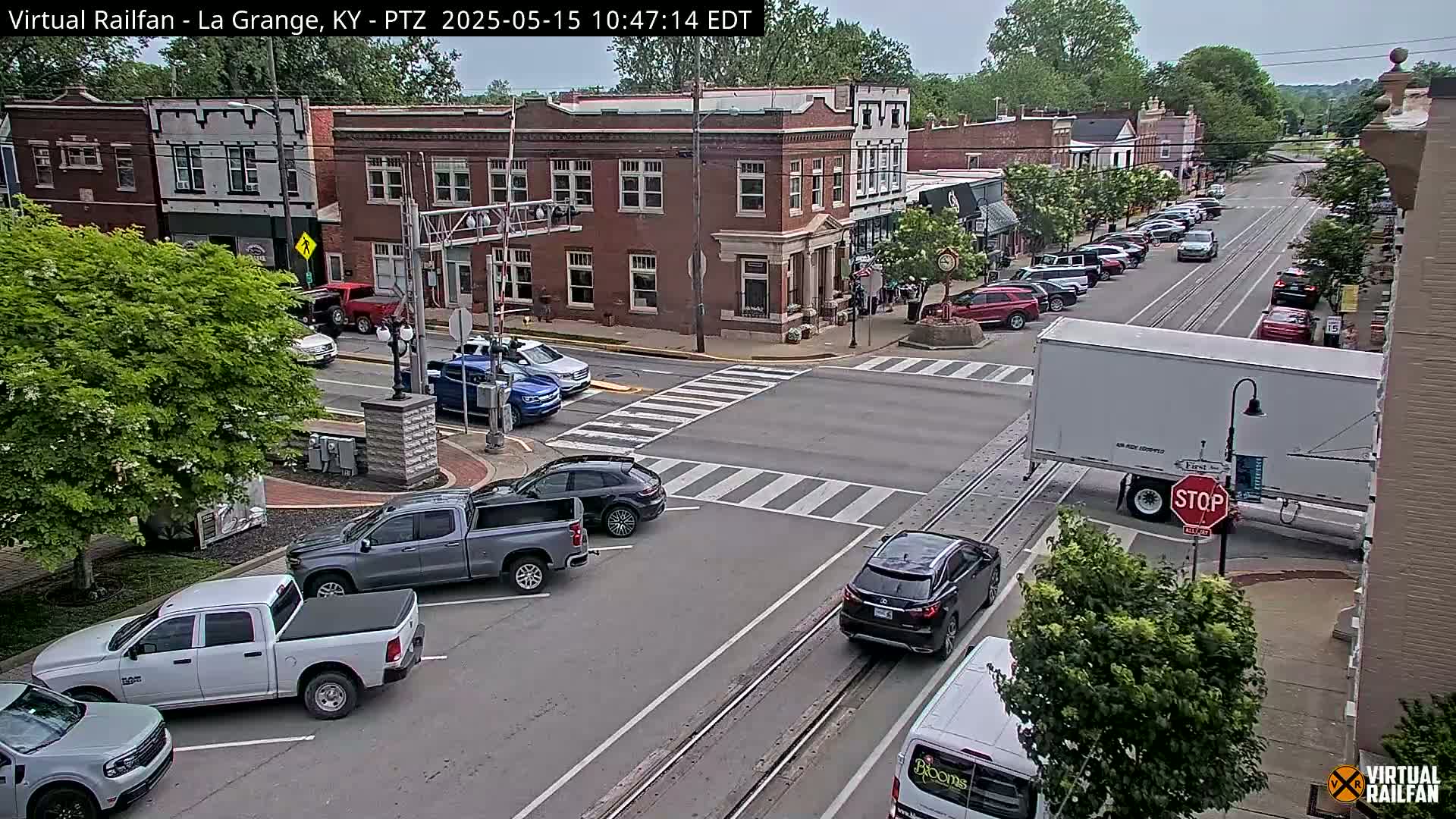 A mostly sunny day shows a town square with several cars and trucks parked and driving, including a large white semi-trailer truck stopped at a crosswalk near brick buildings and lush green trees.