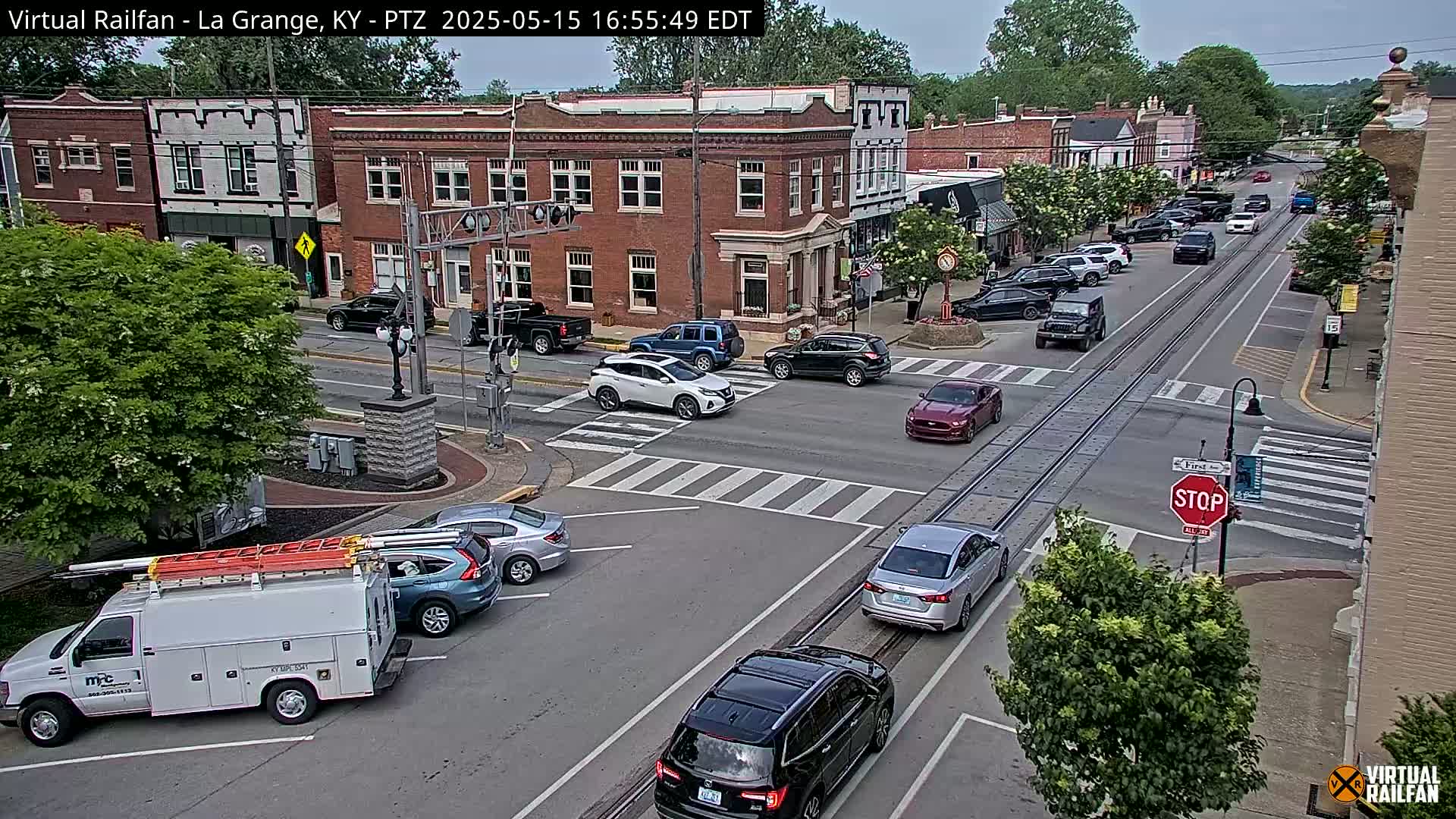 A partly sunny street scene shows a multi-lane road with cars, trucks, and an intersection; brick buildings line the street, and lush green trees are visible.