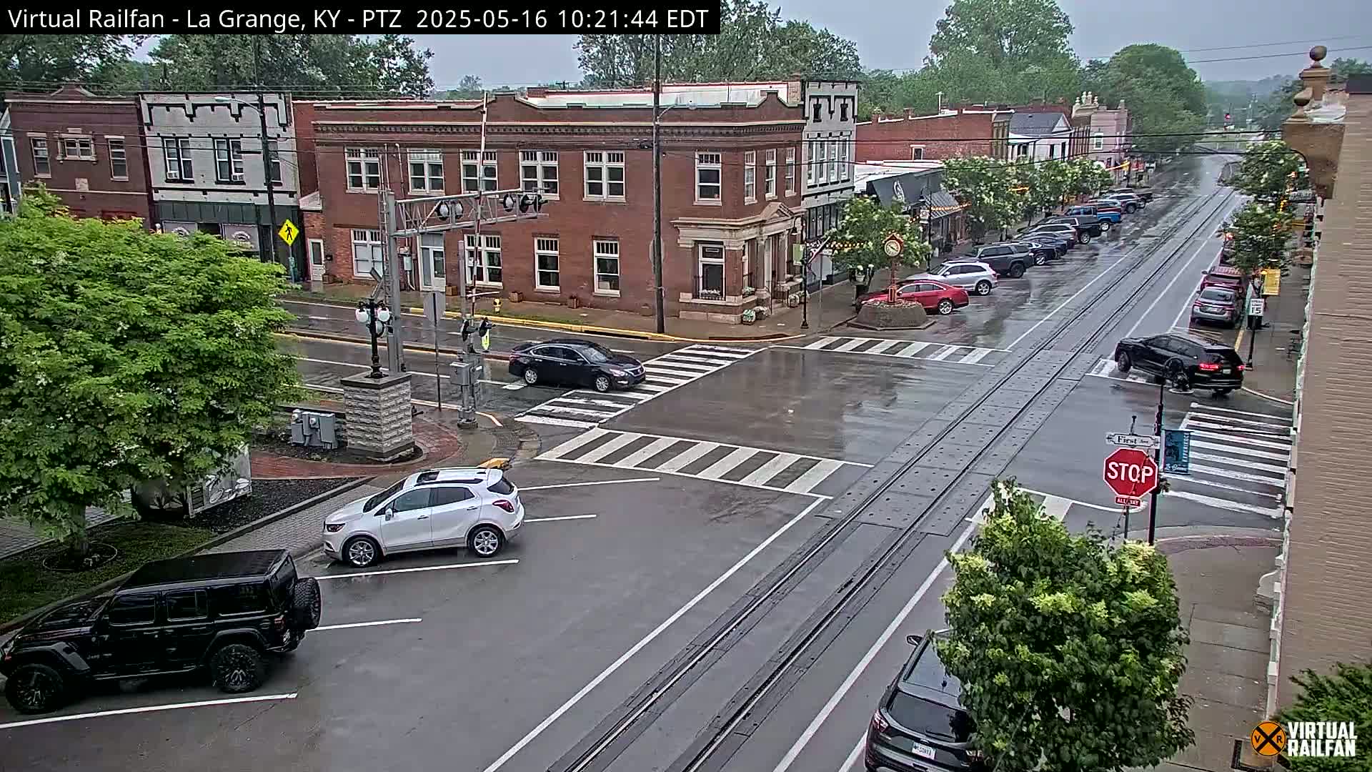 A rain-slicked street scene in a town features several cars at a crosswalk and parked along the street, with brick buildings and lush green trees lining the road.