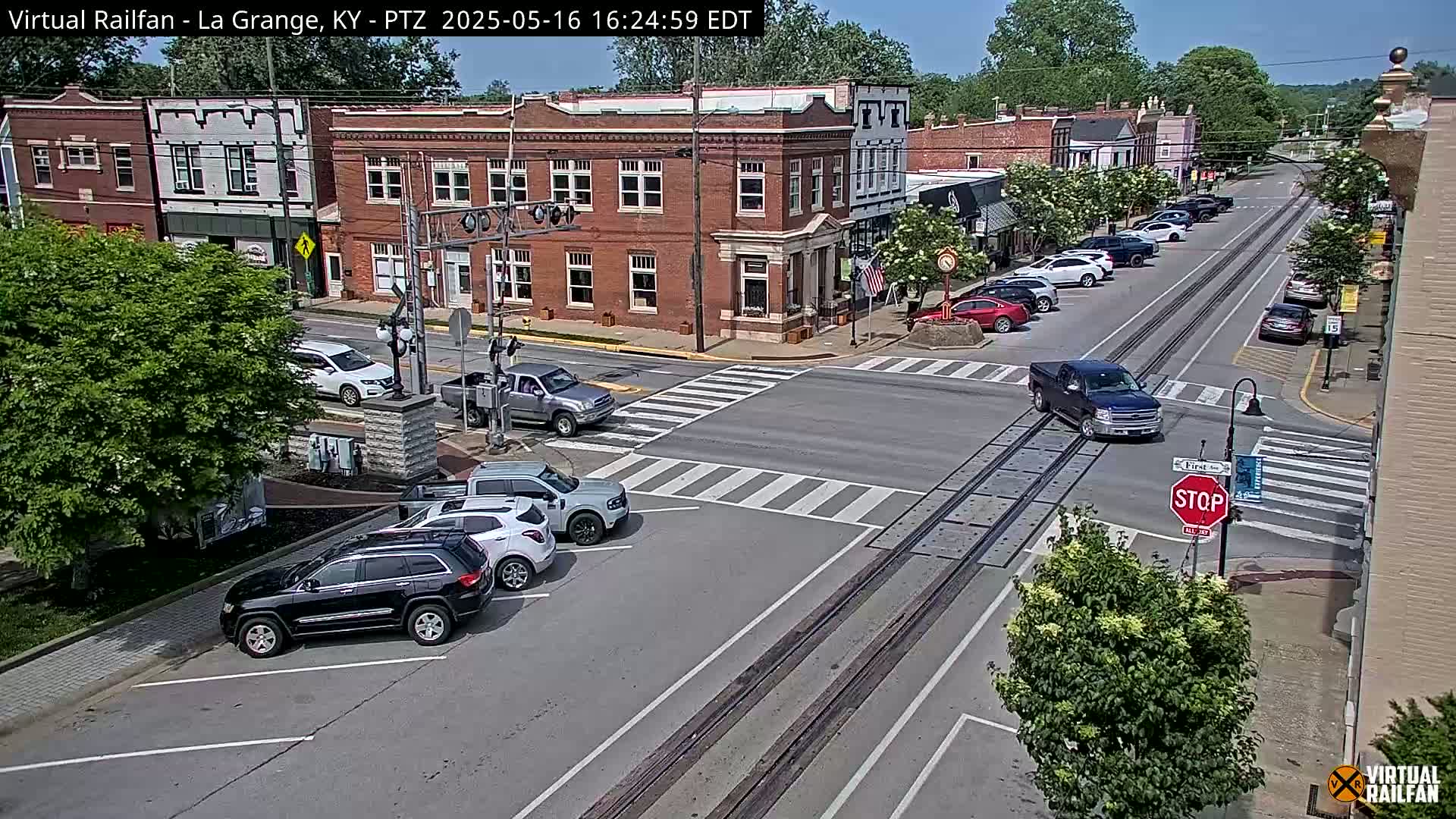 A sunny, daytime street scene shows a town square with brick buildings, parked cars, and a few vehicles driving through an intersection with railroad tracks.
