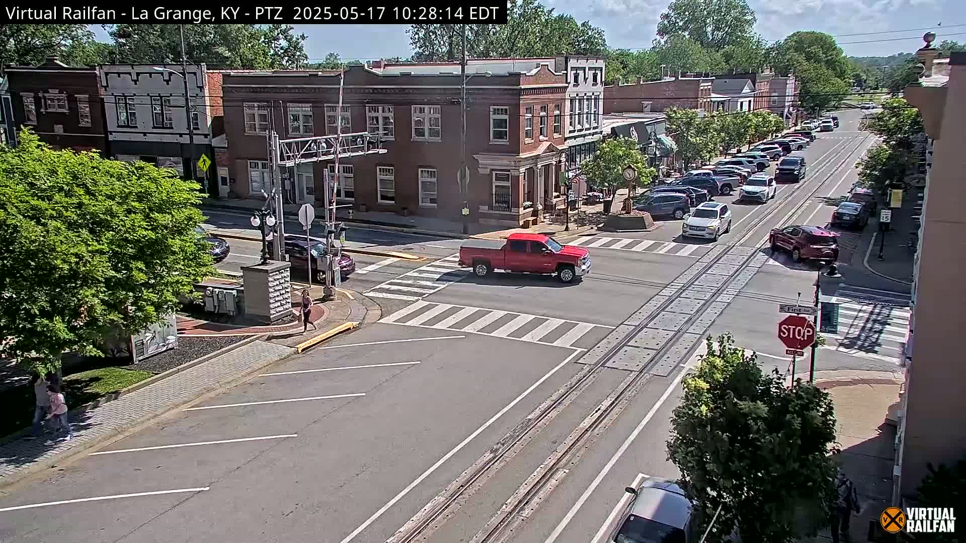A sunny day in a town with brick buildings, parked cars lining the street, a red pickup truck crossing an intersection with railroad tracks, and pedestrians walking on the sidewalk.