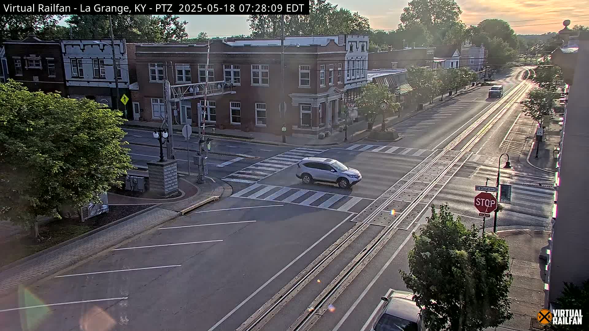 A mostly sunny street scene shows a small town intersection with a few cars and buildings in the early morning.