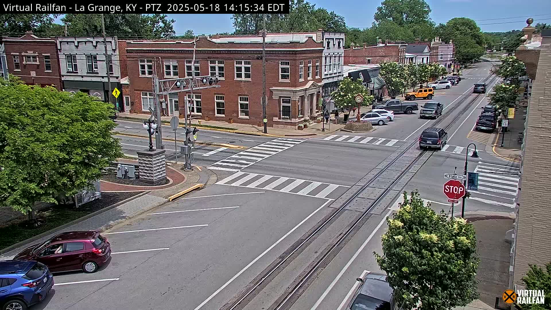 A sunny, tree-lined street scene shows a town's intersection with several cars and pedestrians, railroad tracks in the road, and brick buildings.