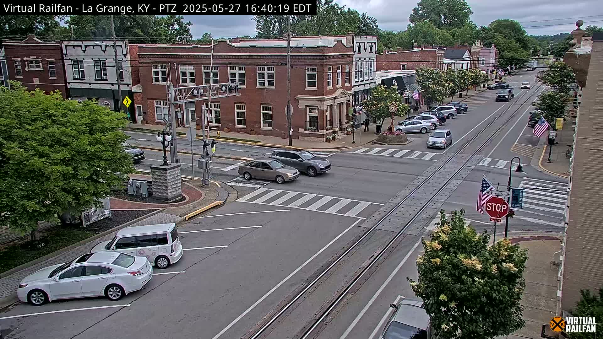 A street scene in a small town on a partly cloudy day shows several cars at an intersection with train tracks running down the middle of the road, and several buildings lining the street.