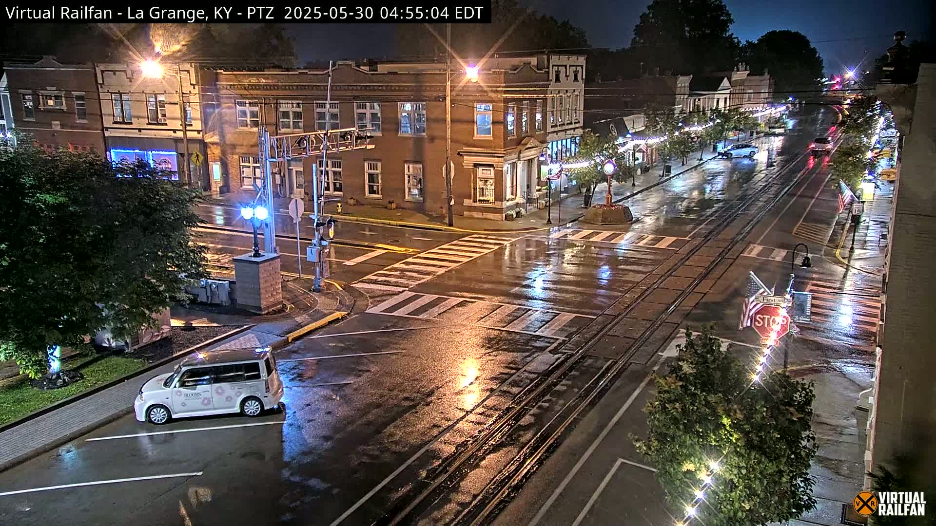 A rain-slicked street scene at night shows a town square with brick buildings, streetlights, and a few cars, including one small white vehicle parked near a corner.