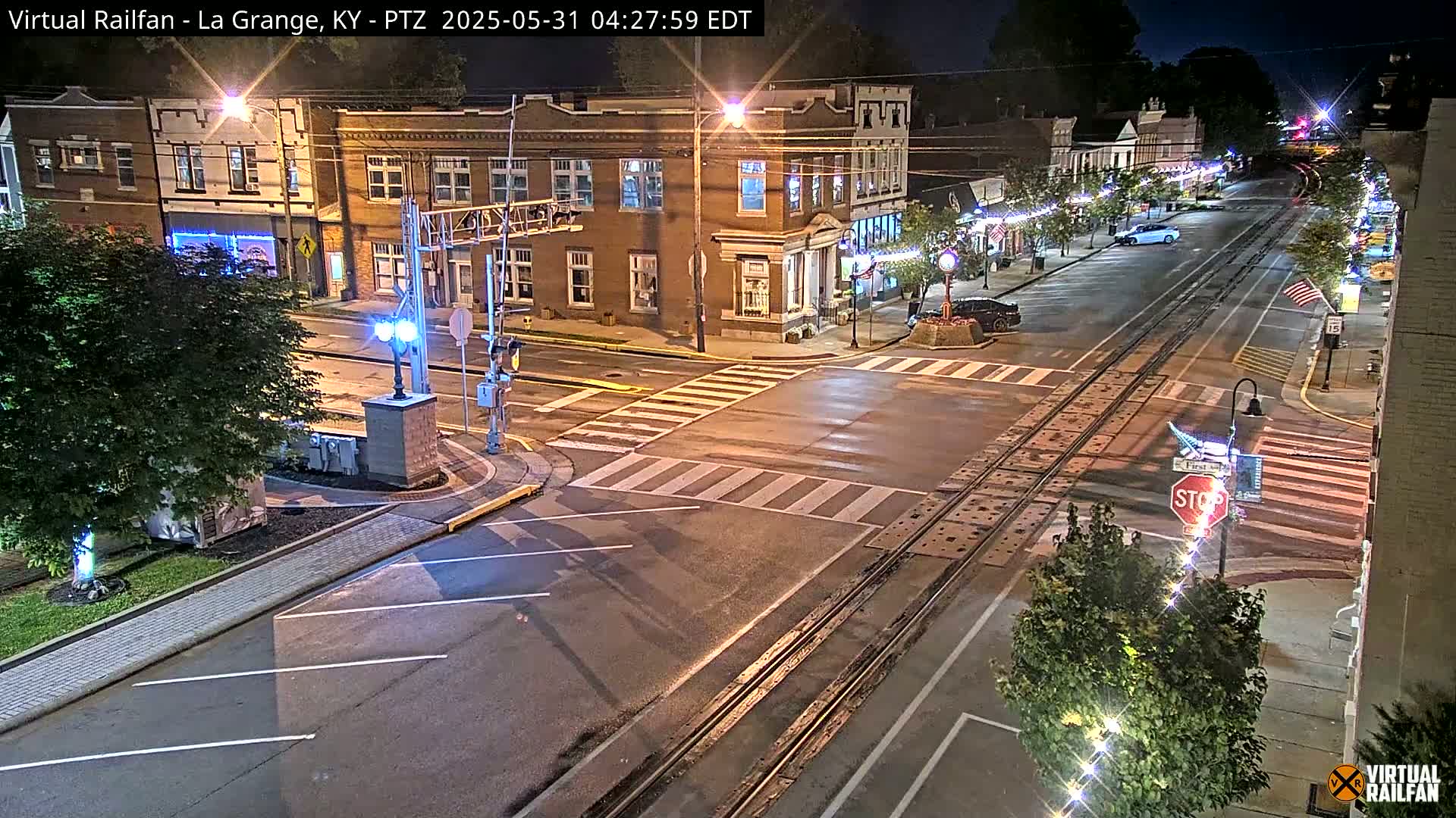 A nighttime view of a town's intersection with train tracks, brick buildings, and streetlights.