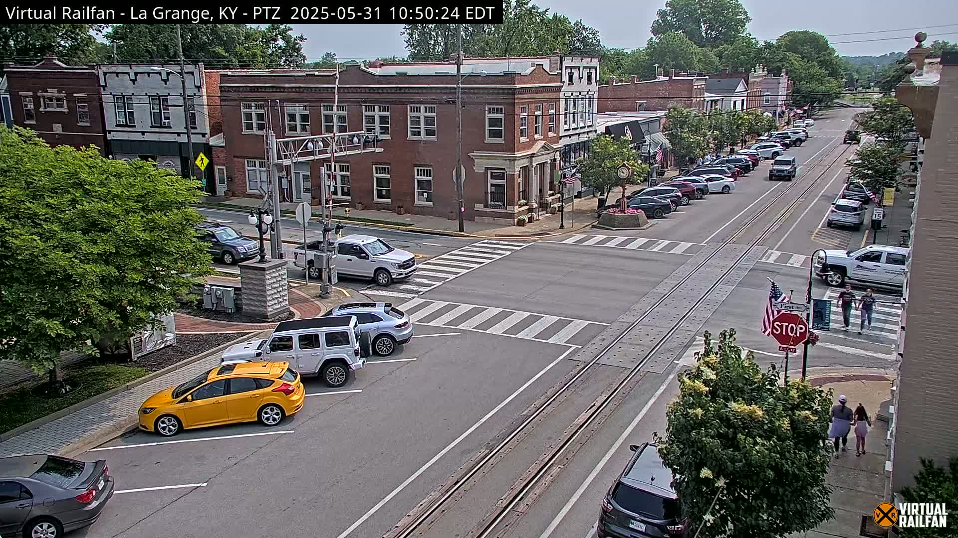 A sunny day in a small town features several cars parked and driving along intersecting streets with train tracks running down the middle, pedestrians walking on the sidewalk, and several brick buildings lining the streets.