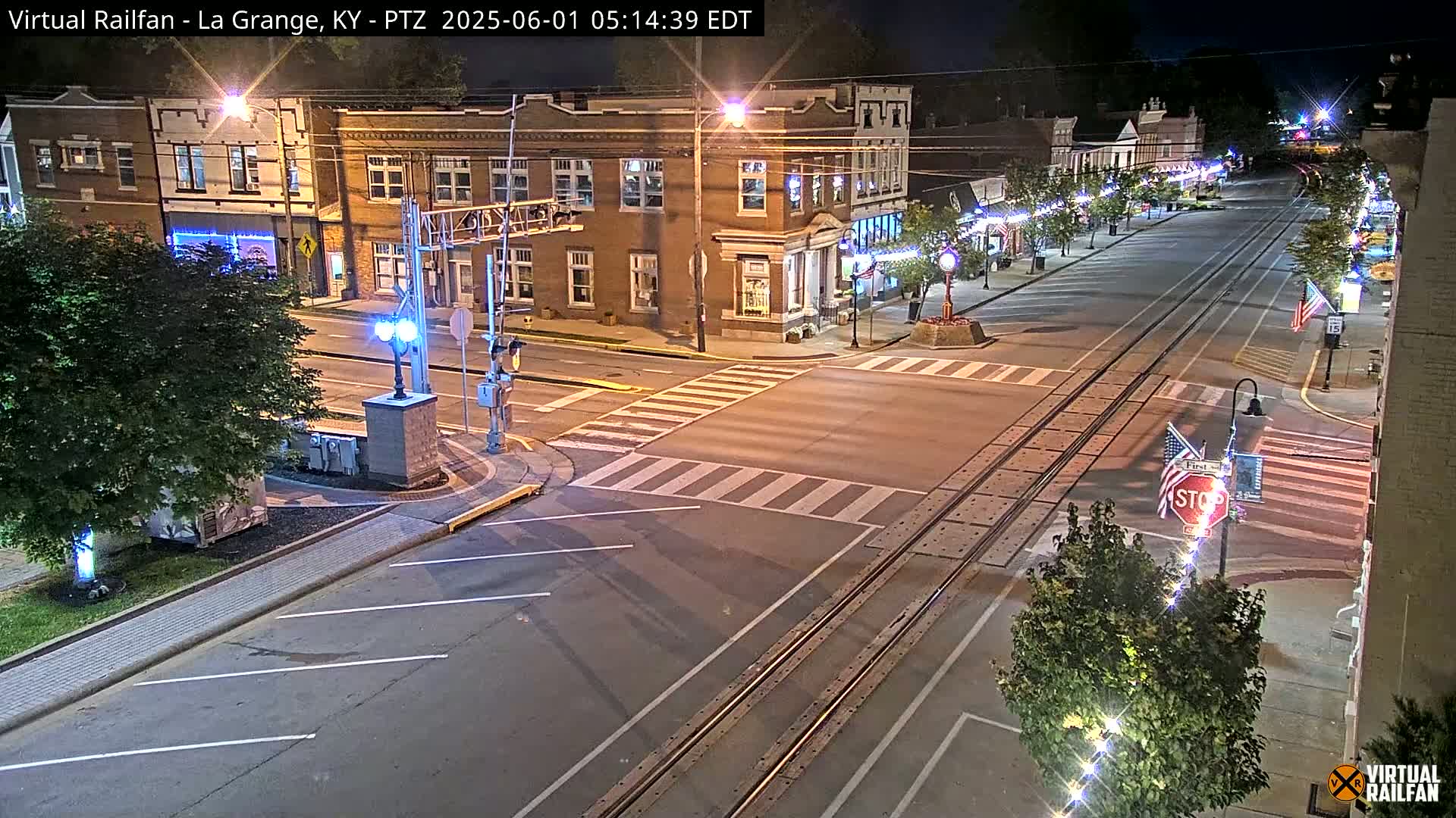 A nighttime street scene shows a brick building and a roadway with train tracks, streetlights, and crosswalks.