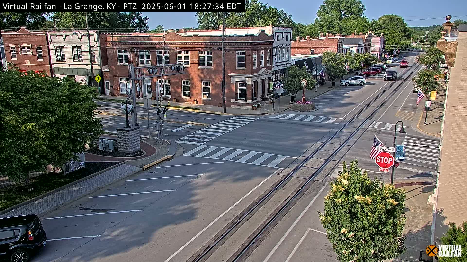 A sunny day in a small town features a street intersection with train tracks running down the center, brick buildings lining the street, and a few cars visible.