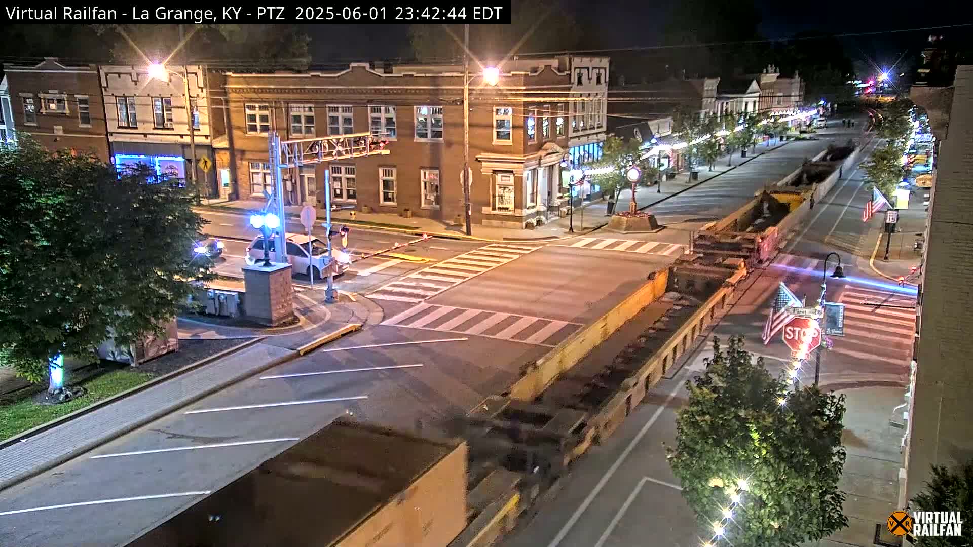 A nighttime street scene shows a town square with a few cars and a large flatbed truck carrying a long load, under mostly clear skies.