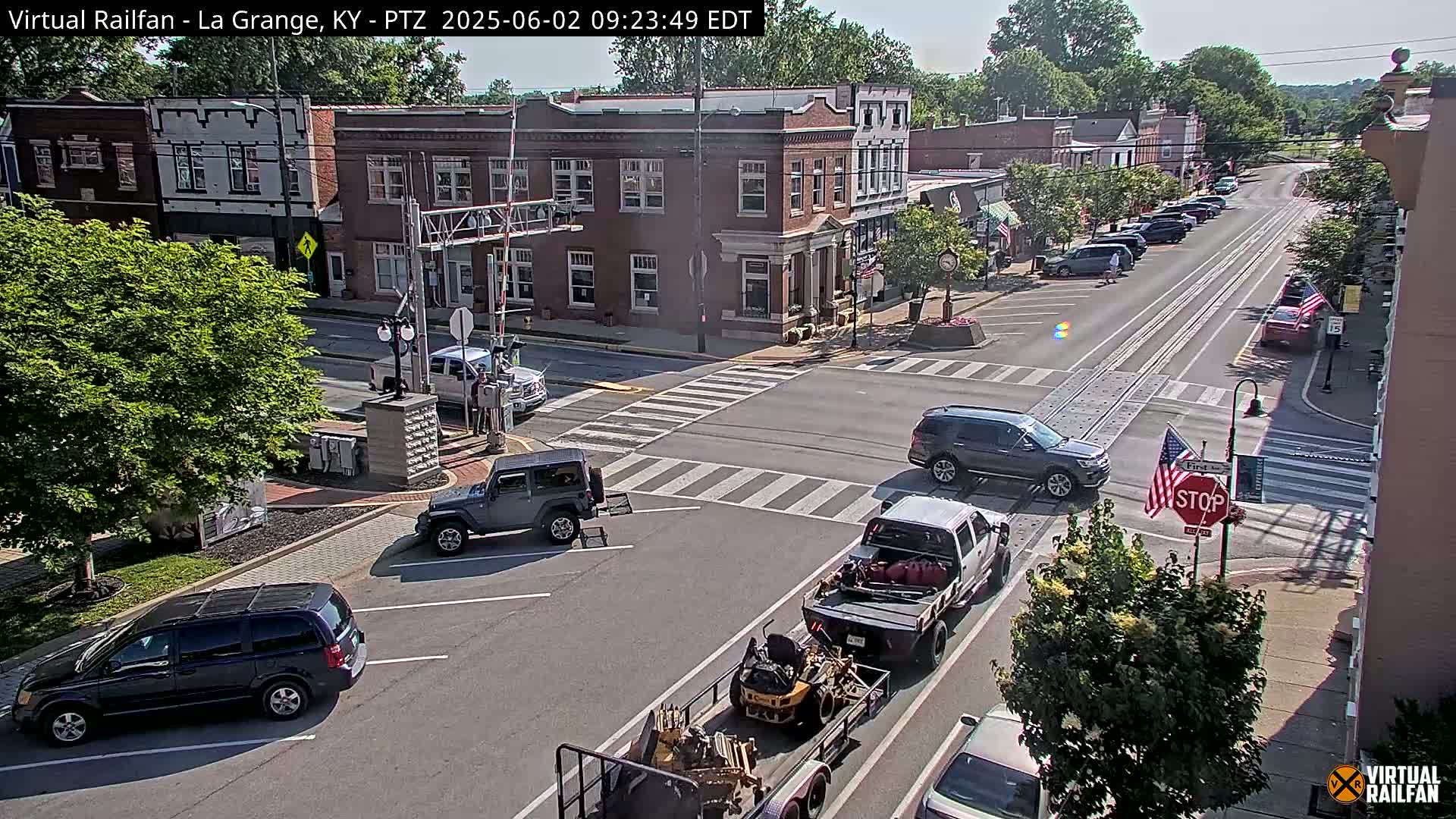 A sunny day in a small town, showing a street intersection with several vehicles, including a Jeep, an SUV, and a pickup truck towing a trailer.