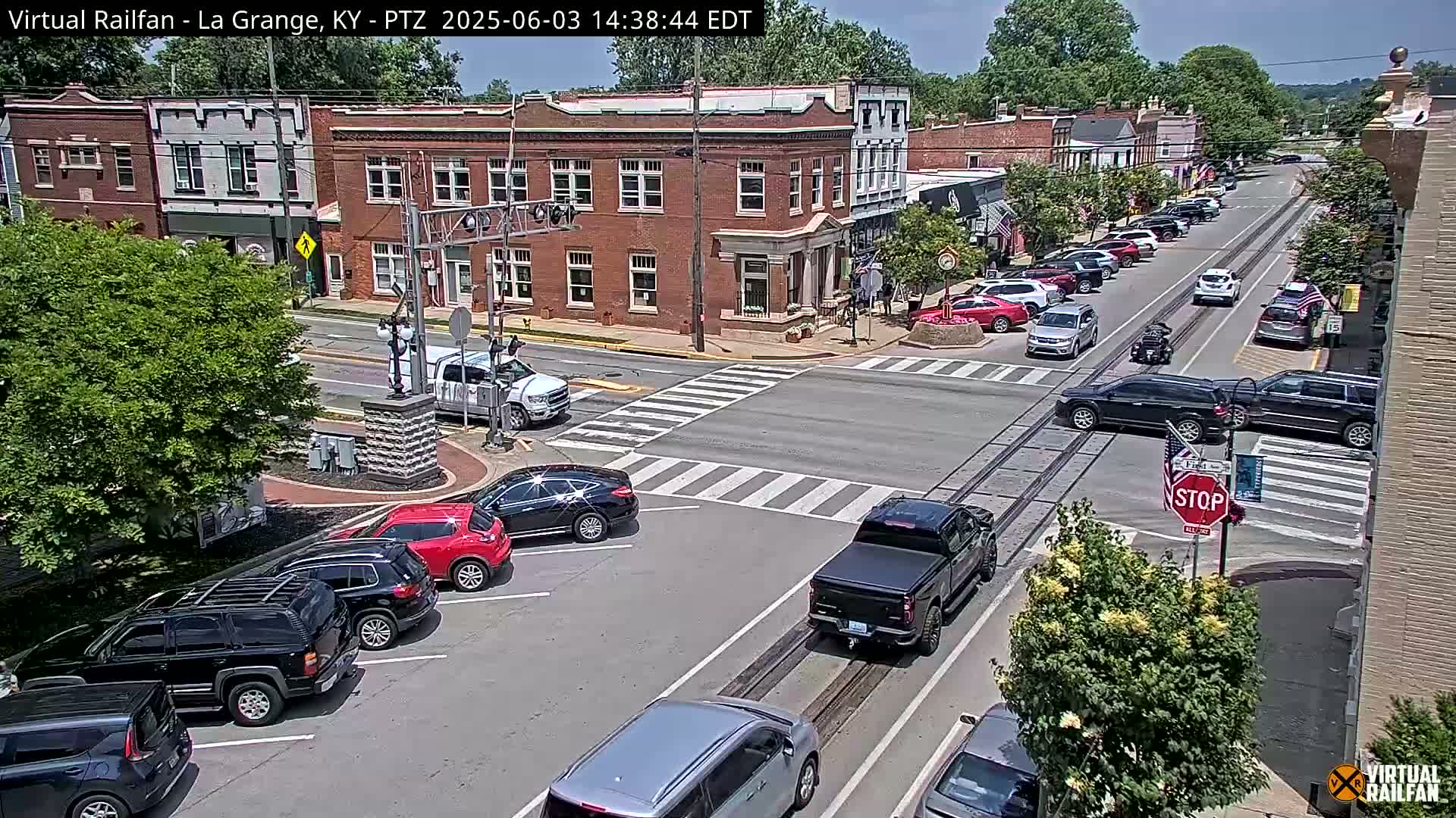 A sunny day shows a town intersection with brick buildings, parked cars, and vehicles traveling on the street and railroad tracks.
