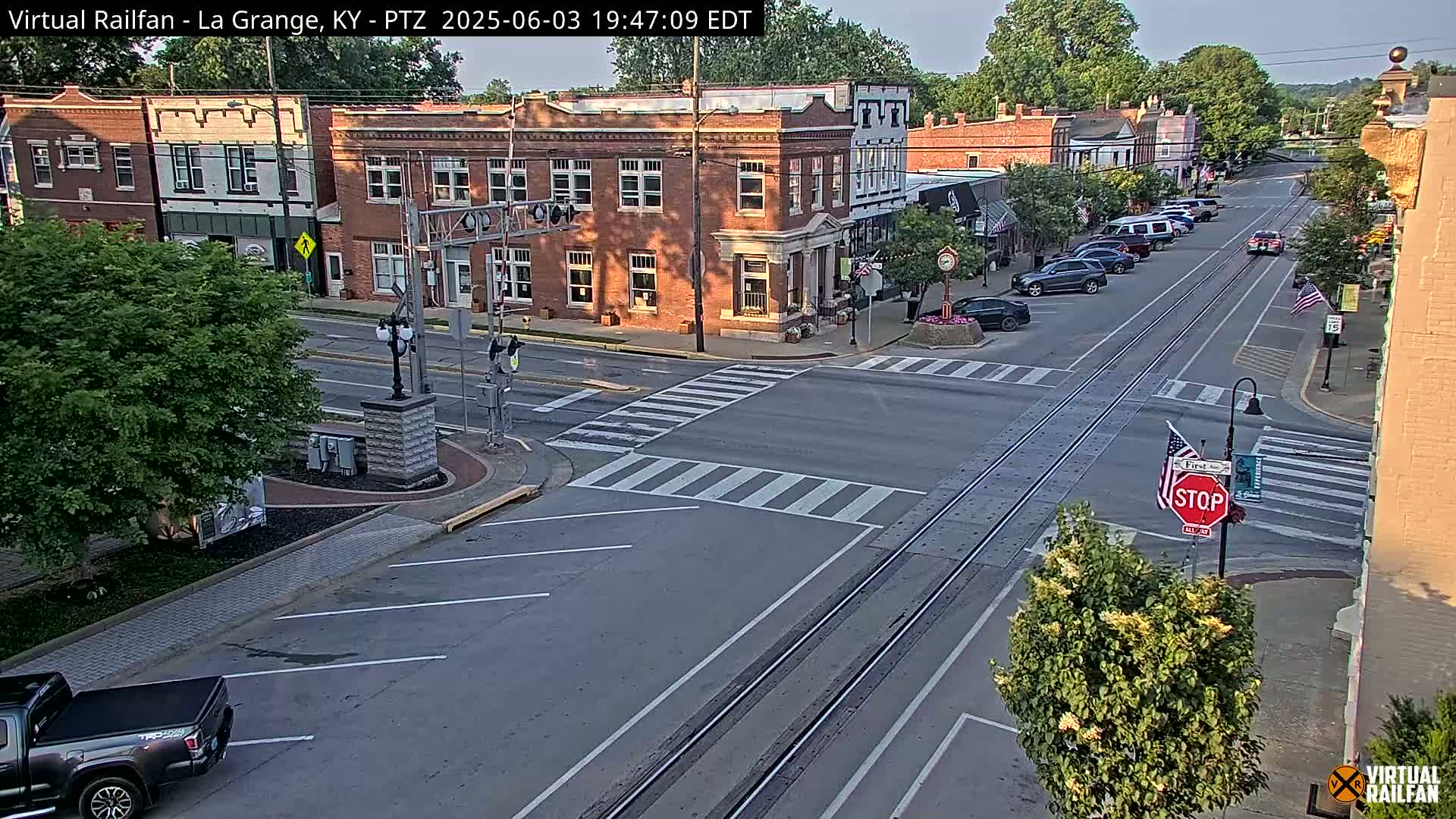 A street scene in a small town on a partly sunny day shows a brick-building lined street with cars parked along the side, a stop sign, and train tracks in the middle of the road.
