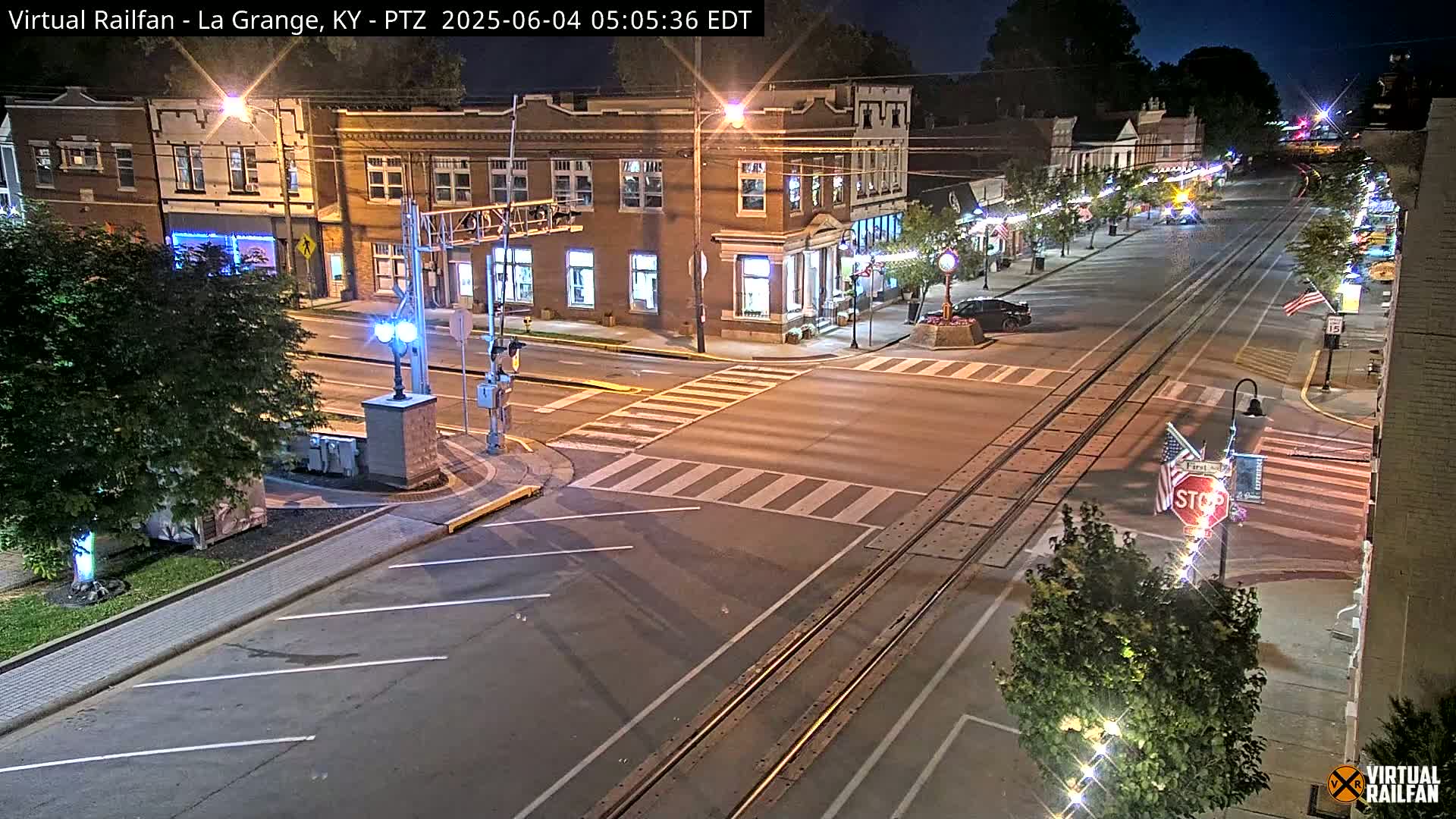 A nighttime view of a town's street intersection with train tracks, featuring brick buildings, streetlights, and a few parked cars.