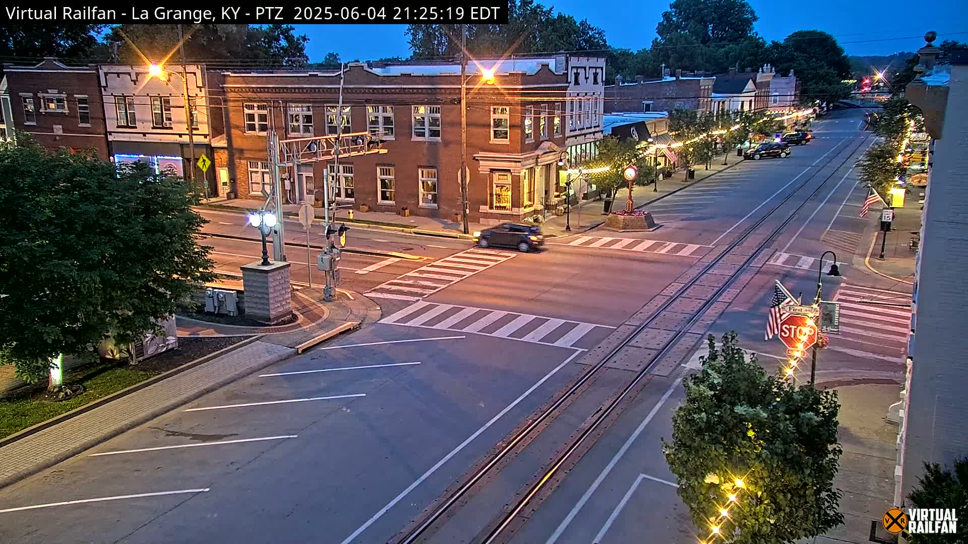 A twilight view of a town's street intersection with brick buildings, streetlights, and train tracks running down the middle of the road.