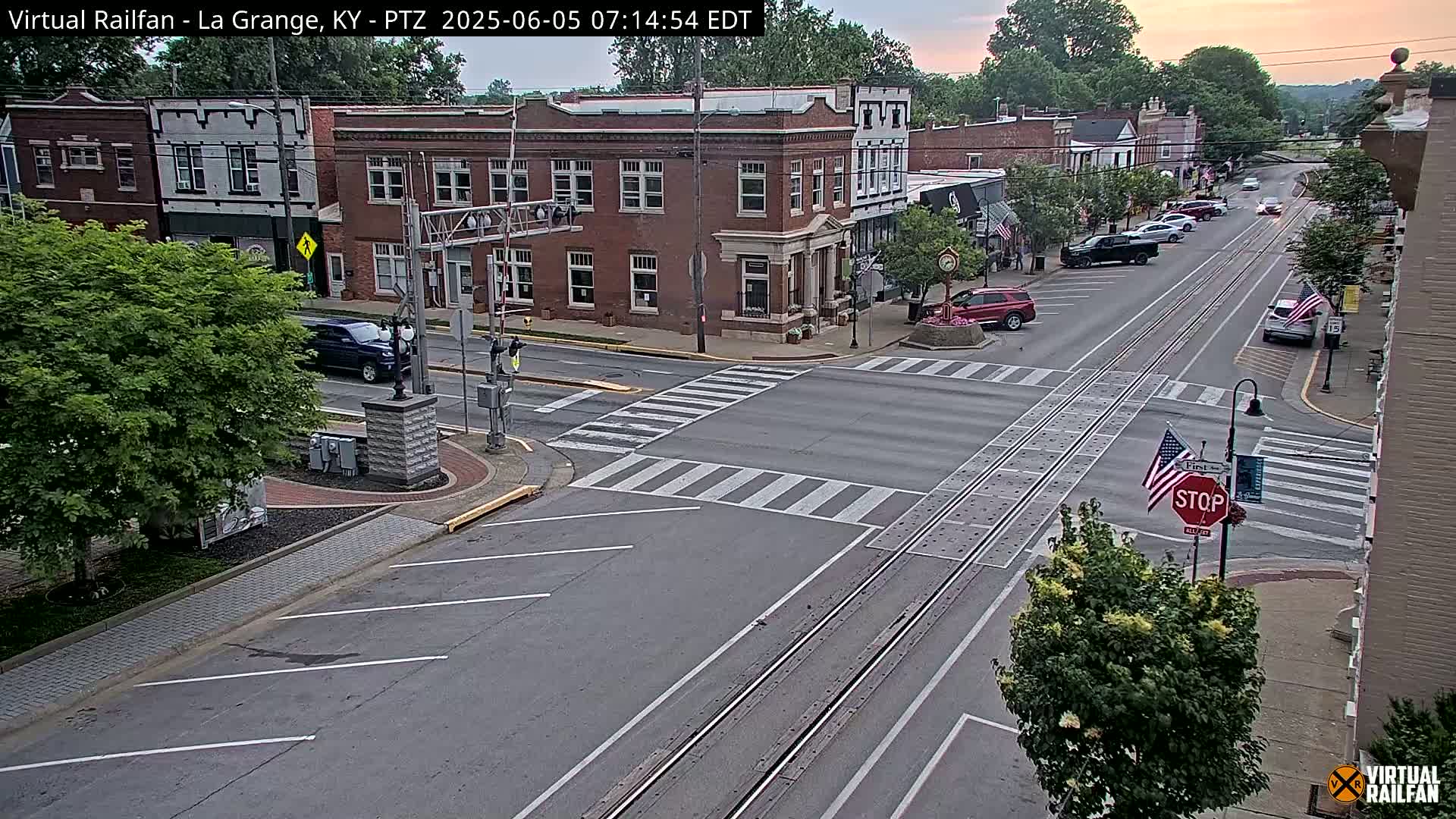 A street intersection in a town features brick buildings, parked cars, and a railway track running down the street, under a mostly clear evening sky.