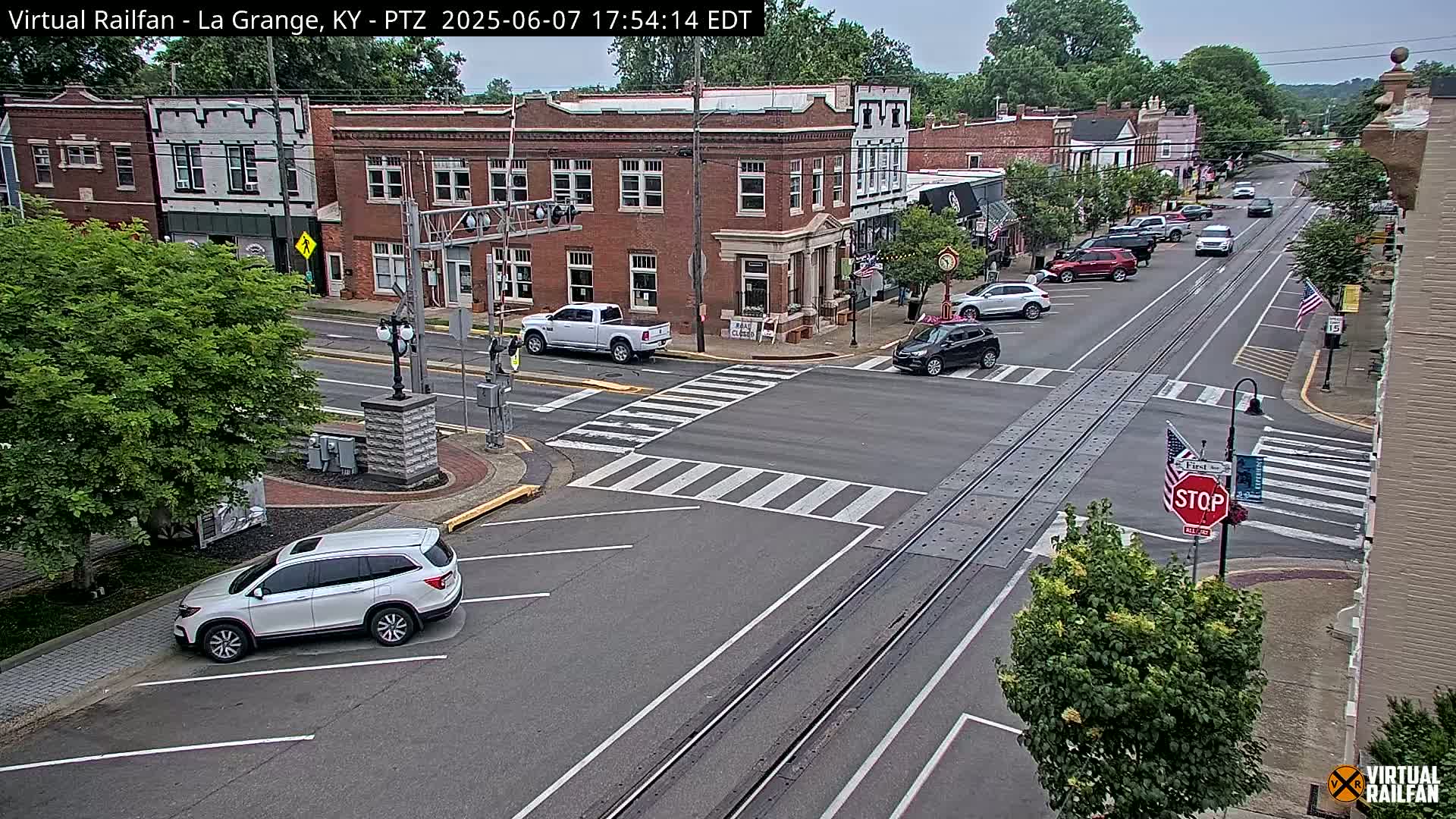 A partly cloudy day in a small town shows a street intersection with several cars, brick buildings, and lush green trees.