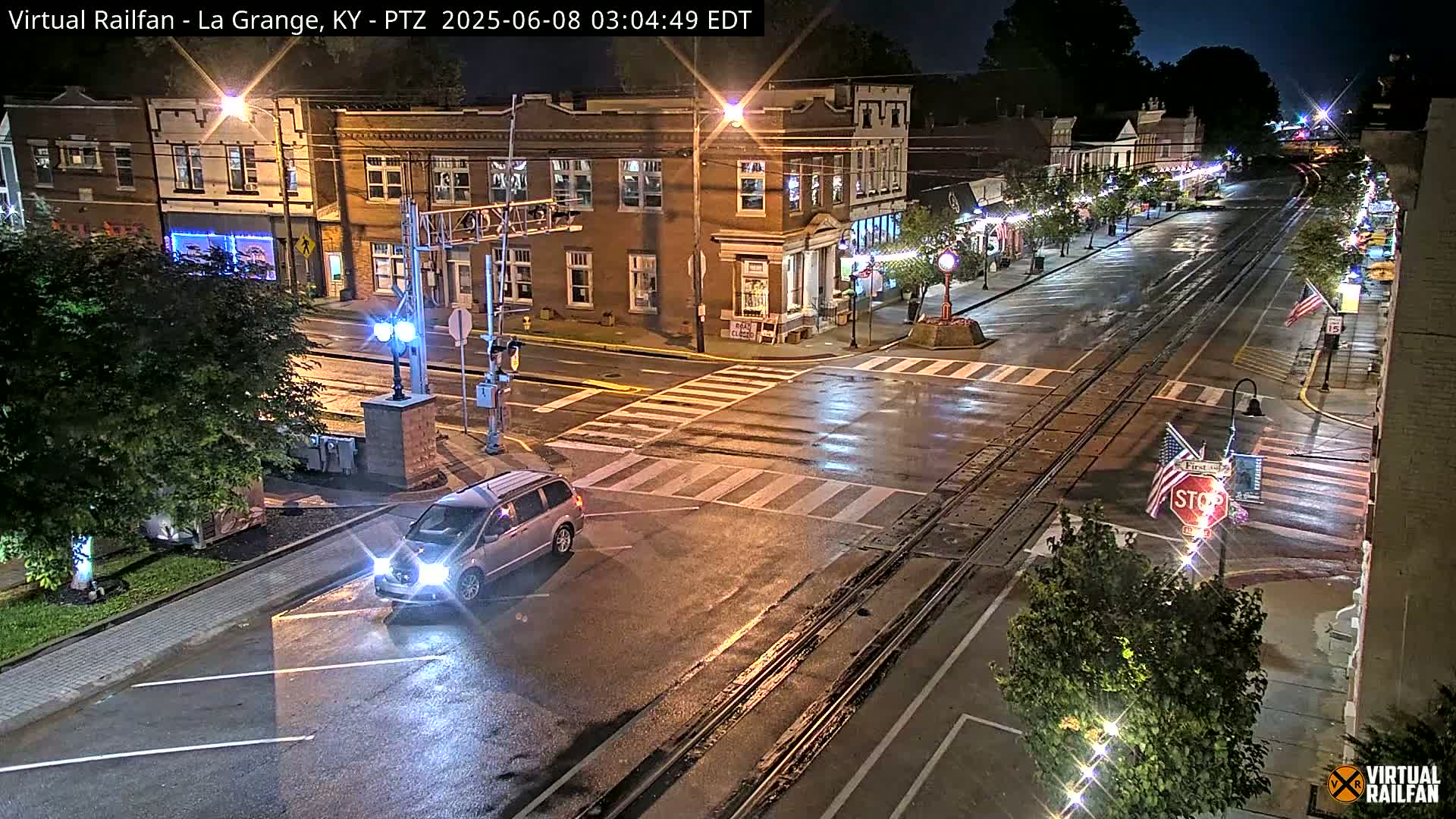 A wet, nighttime street scene shows a minivan at an intersection in front of brick buildings, with train tracks running down the middle of the street.