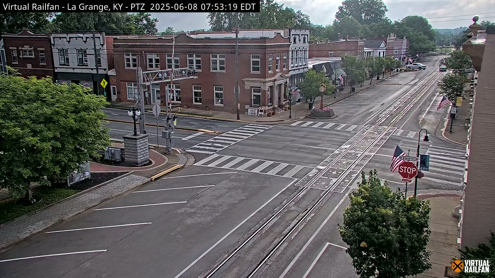 A mostly empty street in a small town, under partly cloudy skies, shows train tracks running down its center and several brick buildings lining the sides.