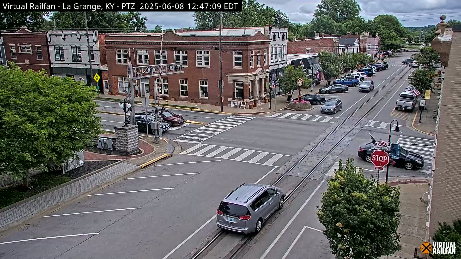 A partly cloudy day in a small town shows several cars at an intersection with brick buildings lining the street and trees along the sides.