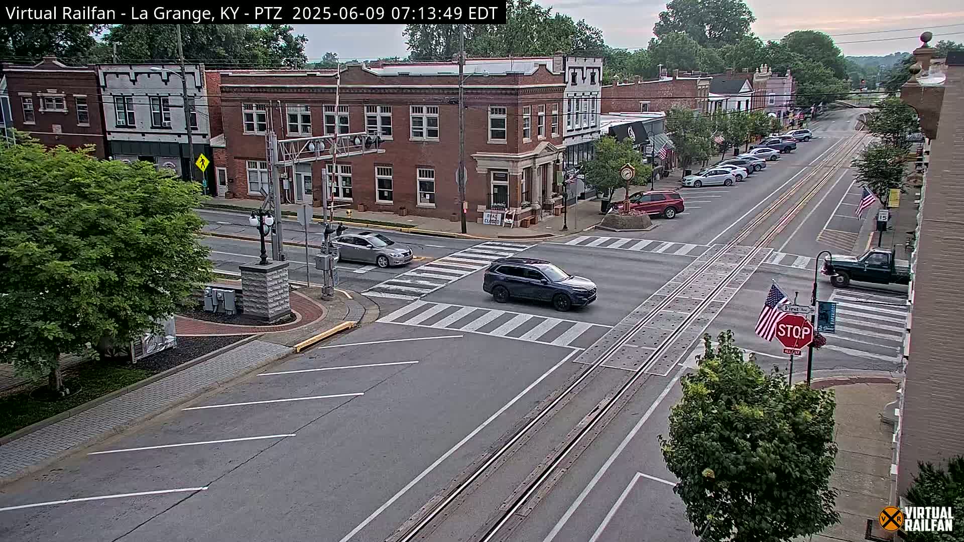 A mostly sunny view of a town's street intersection with cars at a crosswalk, brick buildings lining the street, and several American flags displayed.