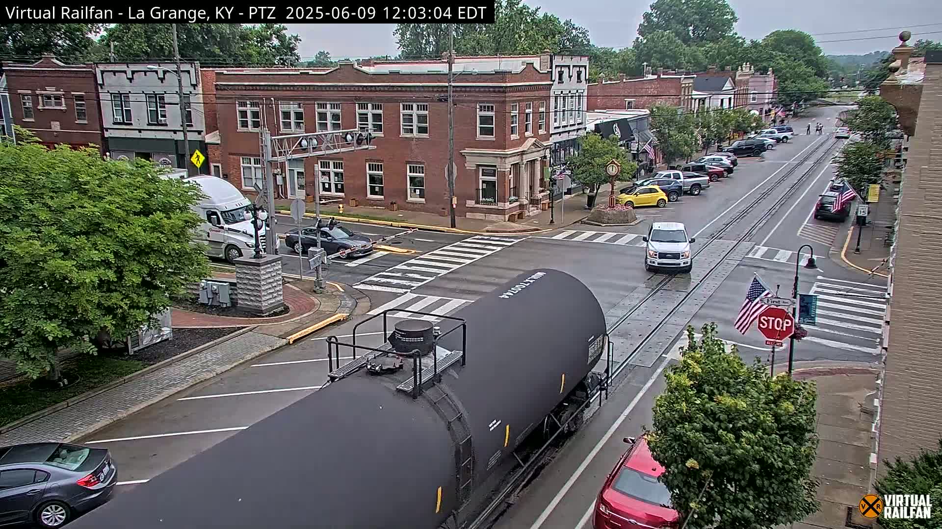 A black railway tanker car is passing through a town intersection on a wet street, with several cars and trucks nearby under an overcast sky.