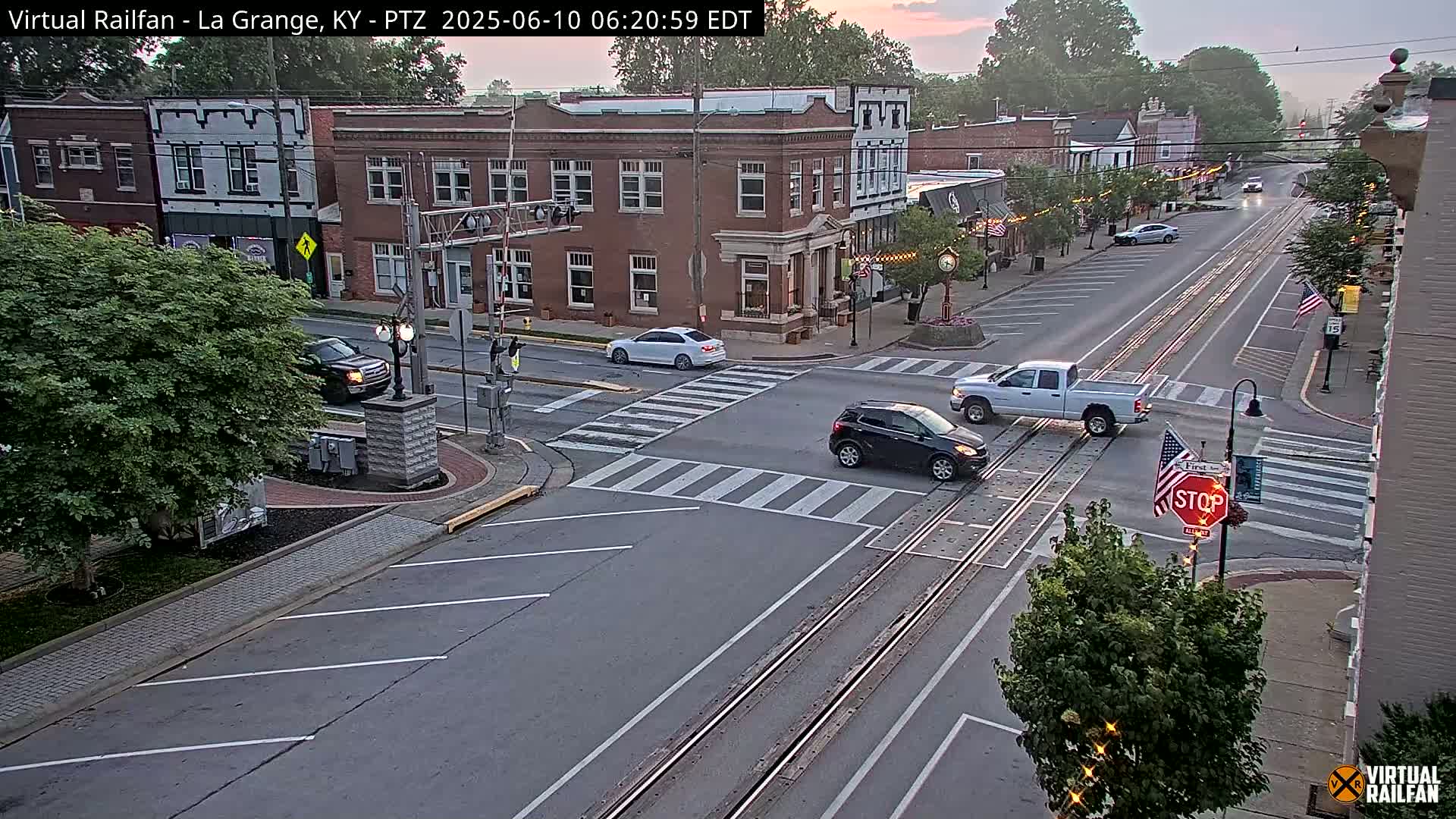 A quiet, overcast morning scene shows a town square with several cars at a railroad crossing, brick buildings lining a street, and lush green trees.